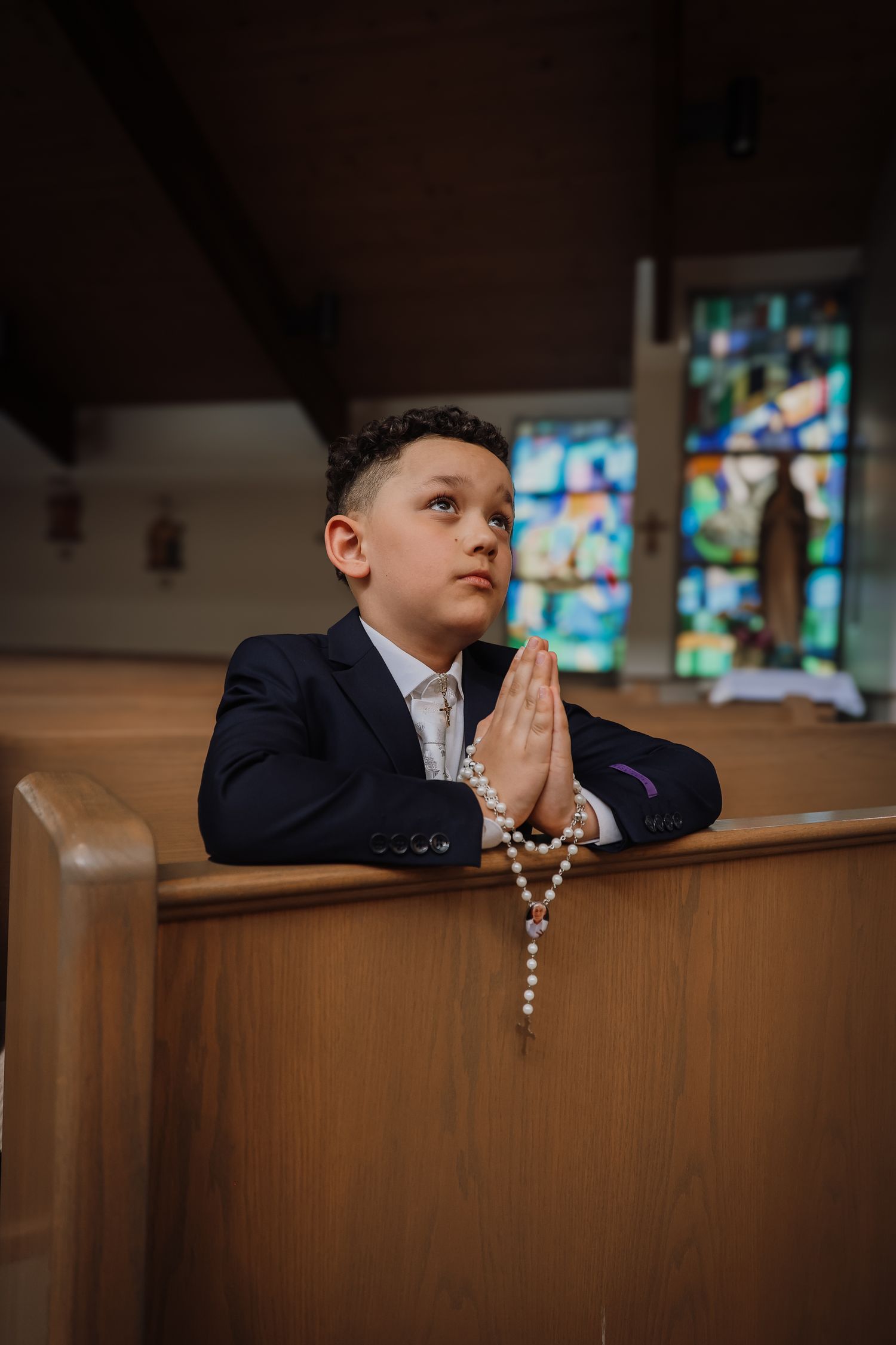 A young boy in a navy suit rests thoughtfully against a wooden church pew while holding rosary beads.