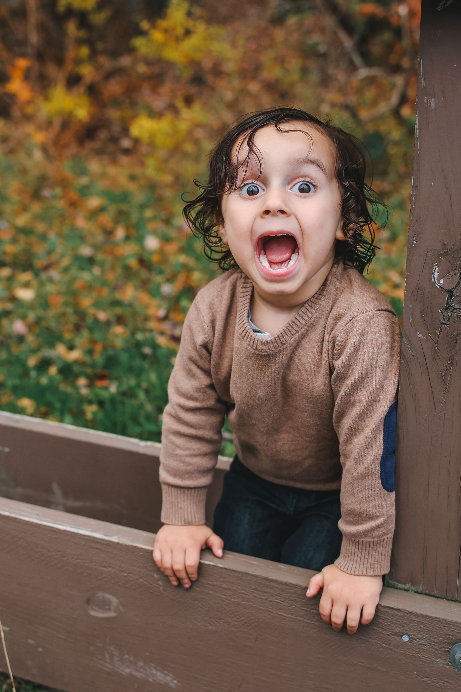 A child in a brown sweater peeks over a wooden railing outdoors with playful expressions in autumn.