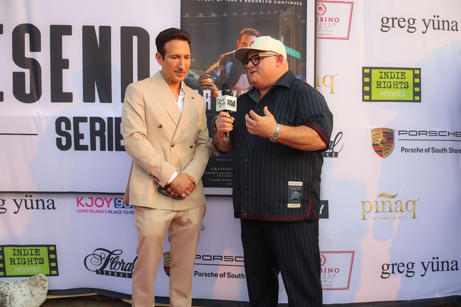 Two people stand together at a media event backdrop with sponsor logos and branding visible behind them.