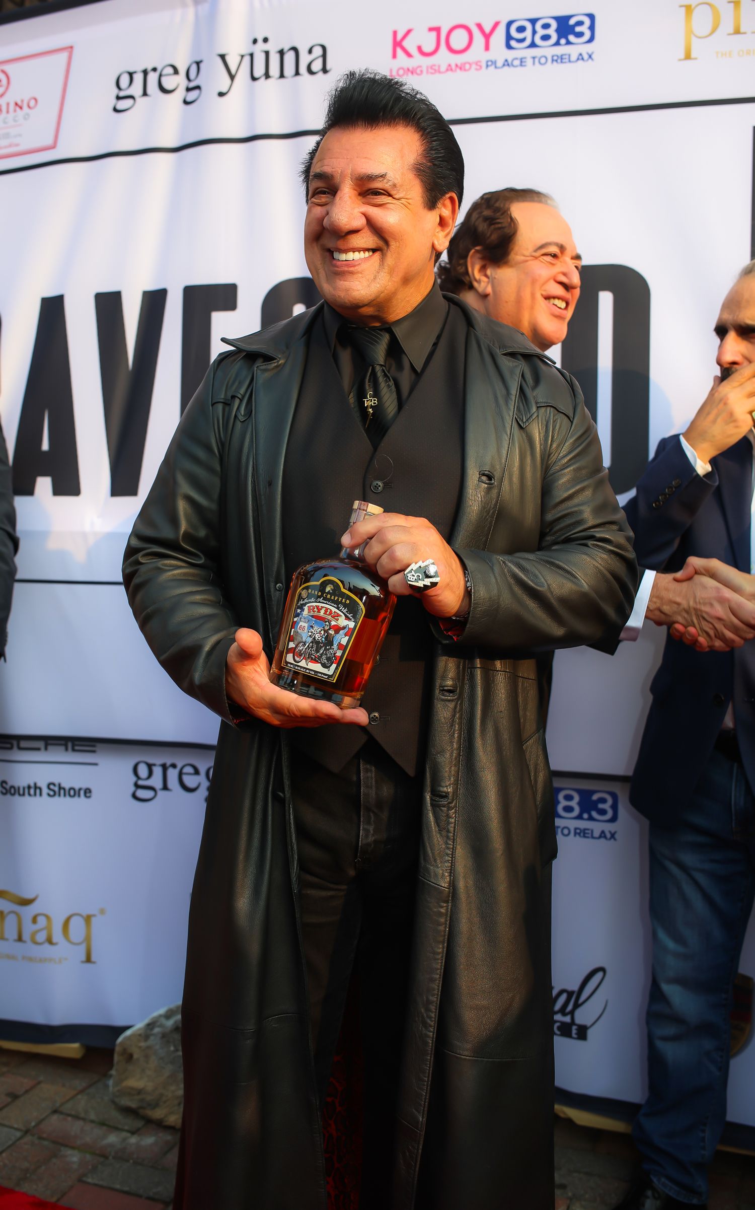 A person in a black coat gestures while holding an award at a media event with KJOY radio station branding visible.