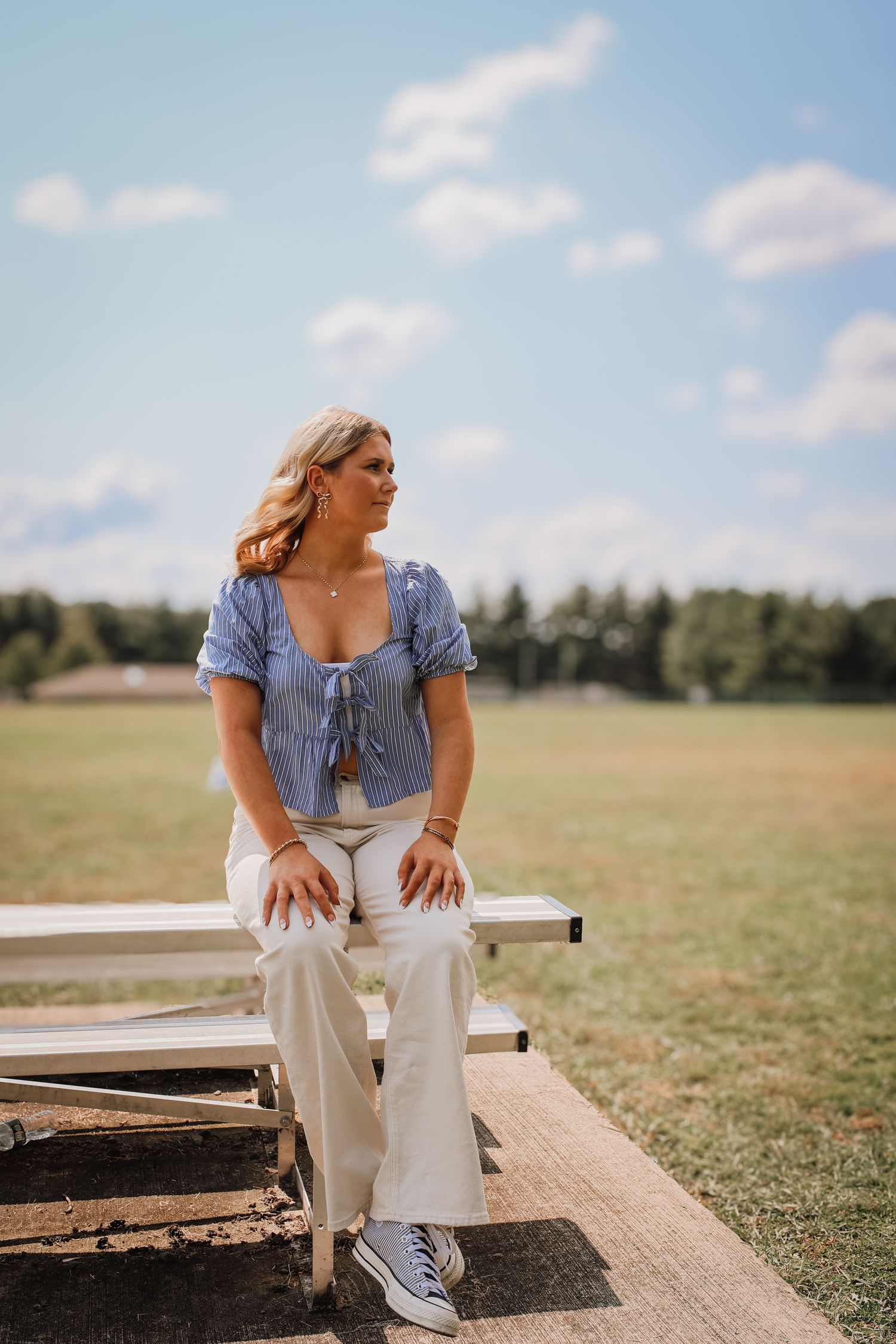 Someone in a blue cropped top and white pants sits casually on a rustic wooden bench in a sunny meadow.
