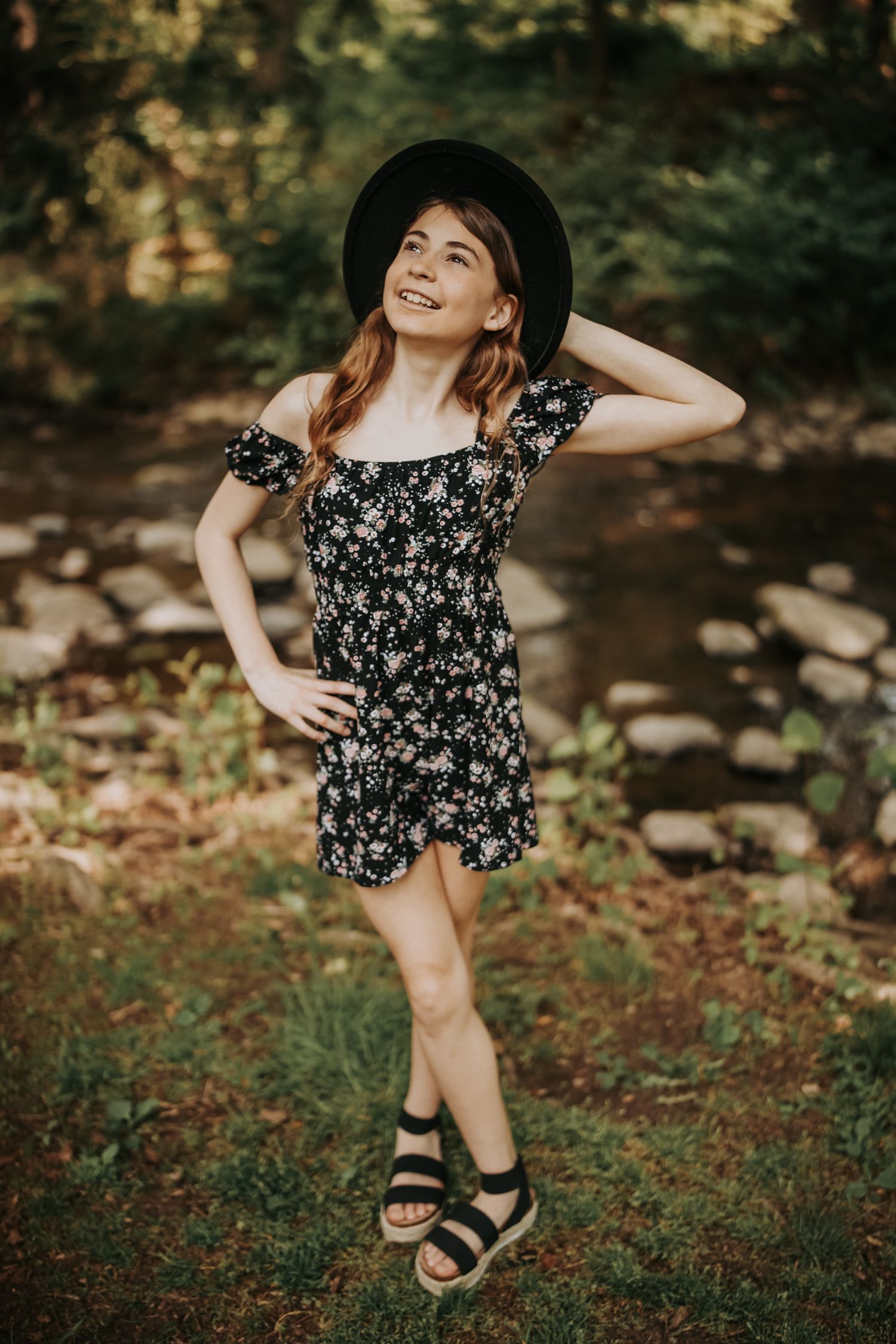 A young child in a floral dress and sandals poses playfully outdoors near a stream and rocks in a natural setting.