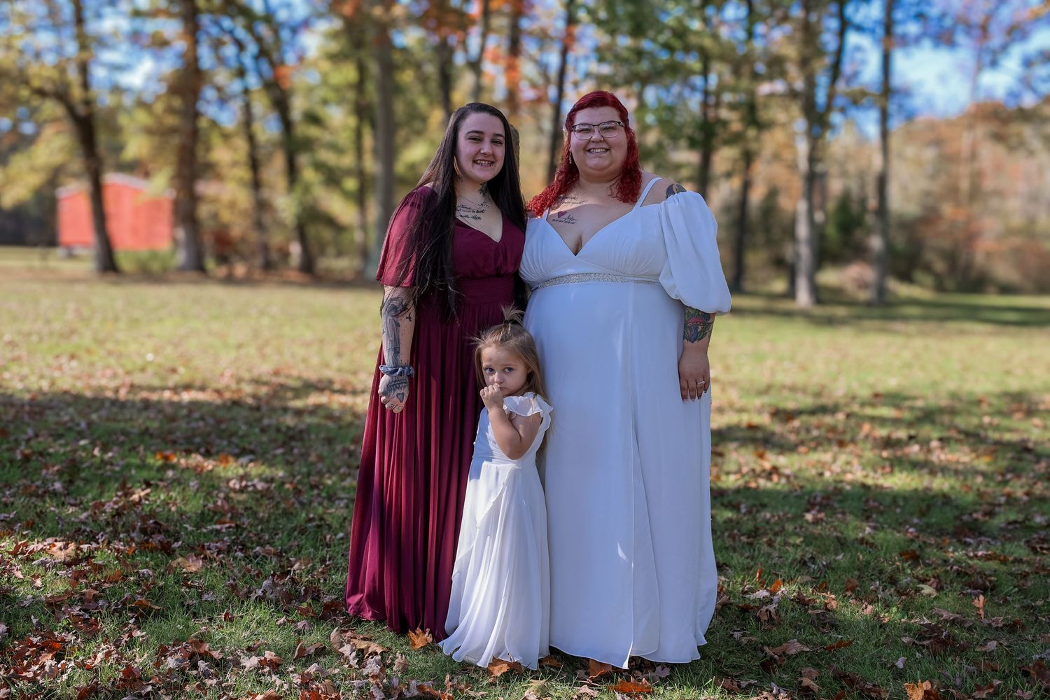 People in coordinating burgundy and white dresses pose together outdoors during autumn with fallen leaves on the ground.