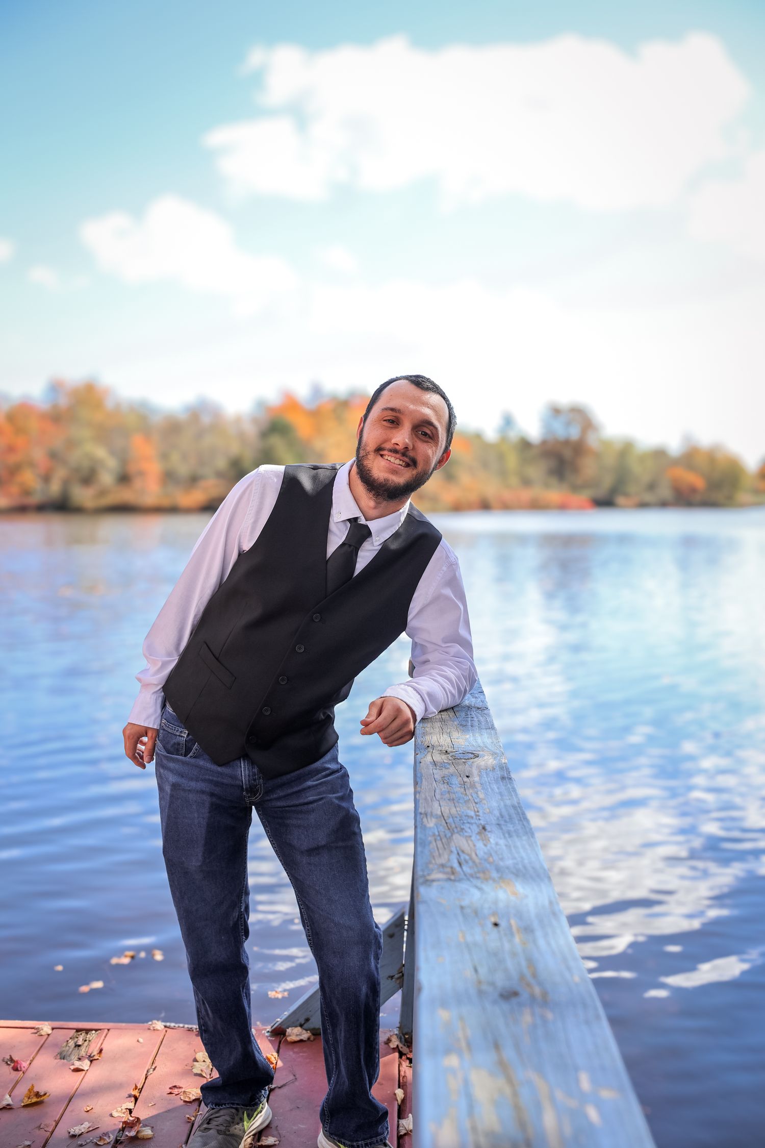 Someone poses casually on a lakeside dock during autumn with colorful fall foliage reflecting in the calm water.