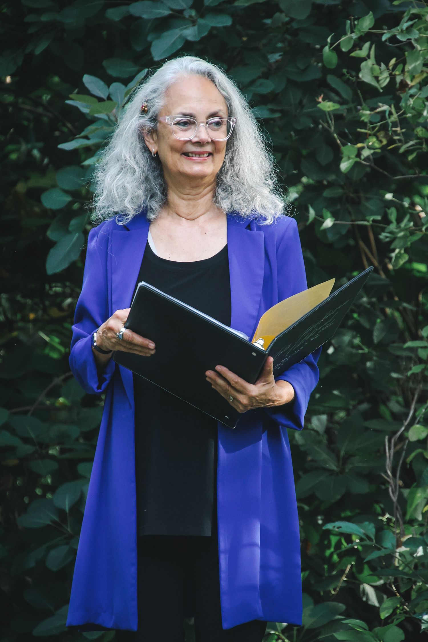 Professional figure in a royal blue blazer and black outfit holding an open folder while standing against green foliage.