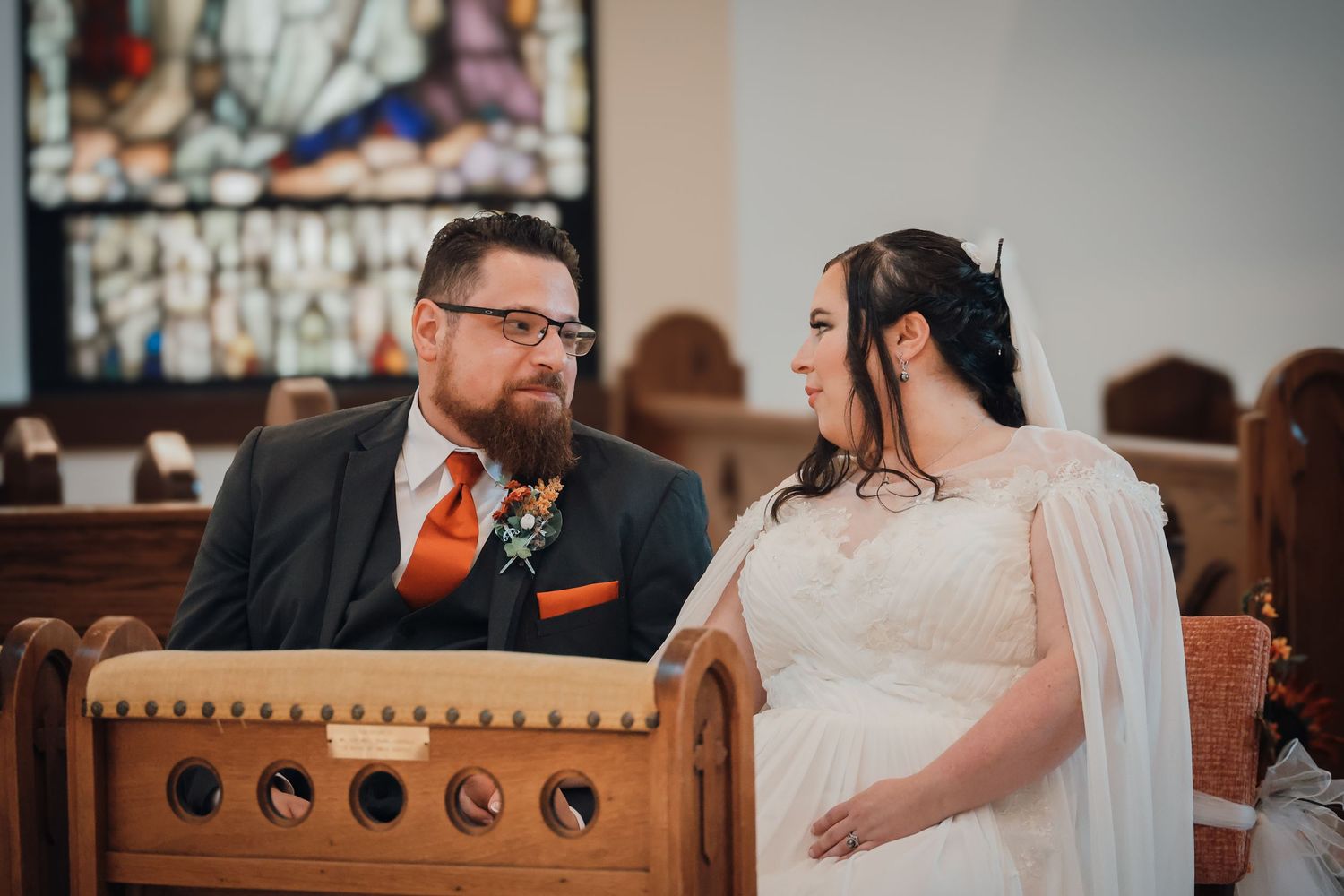 A couple shares a tender moment while seated in a wooden church pew during their wedding ceremony.
