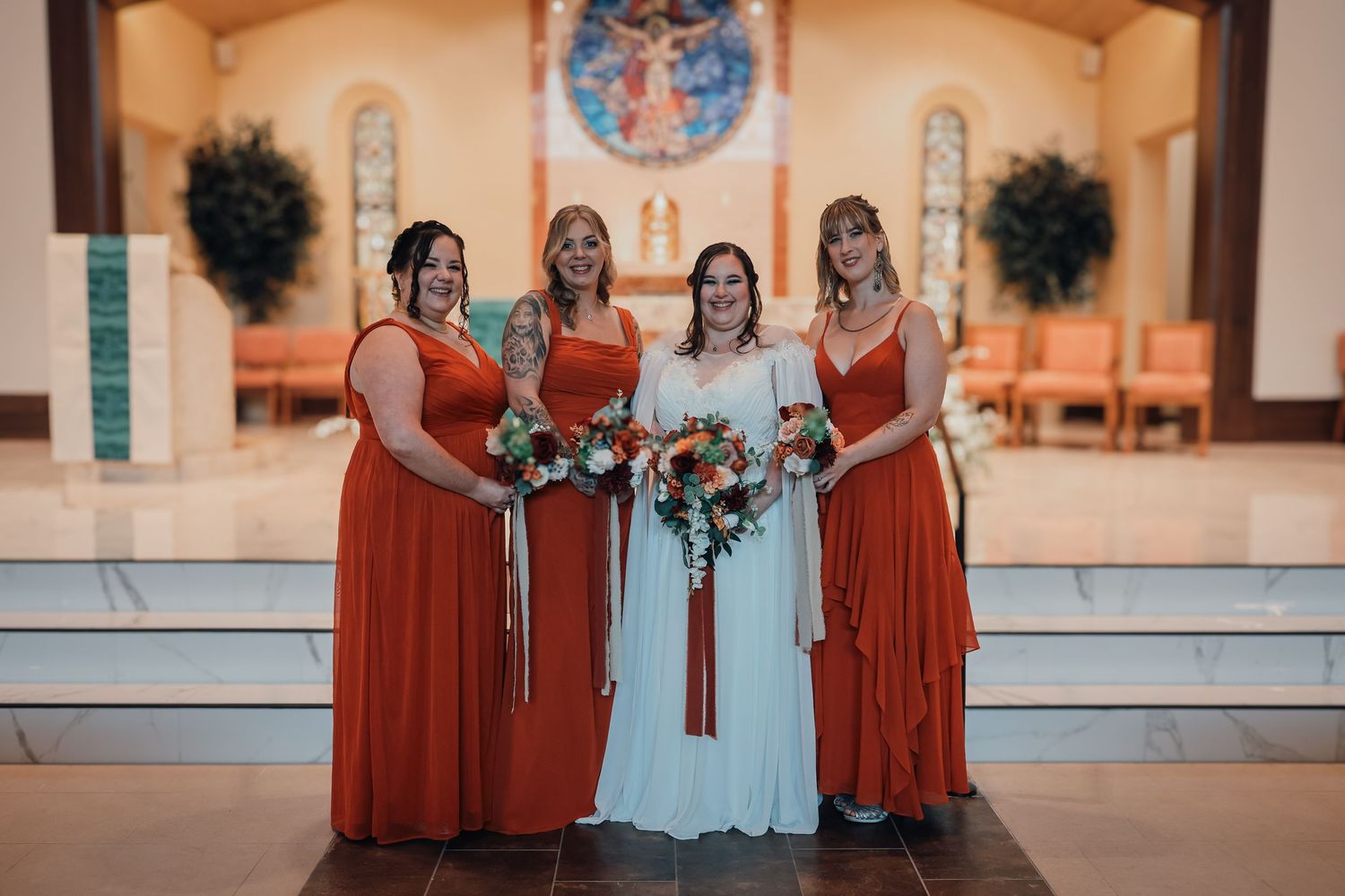 Wedding party wearing burnt orange and white dresses pose together in an elegant church setting.