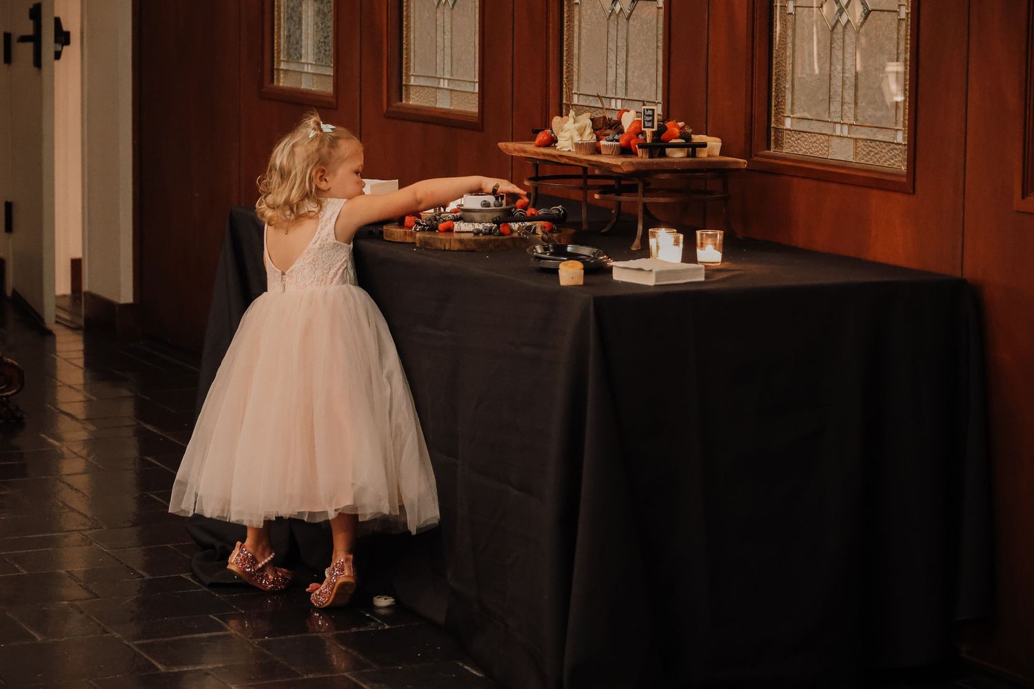 A young guest in a tulle dress reaches for refreshments at an elegant reception table draped in black linens.