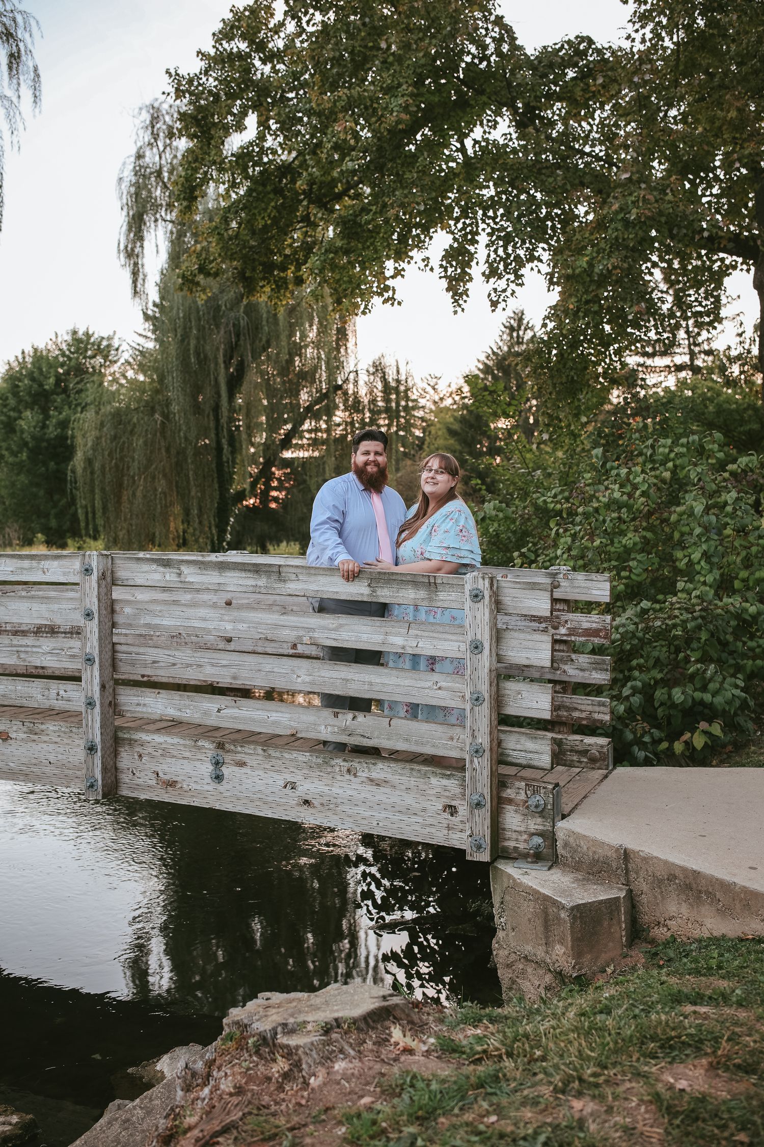 A couple stands together on a rustic wooden bridge over a peaceful stream surrounded by weeping willows at sunset.