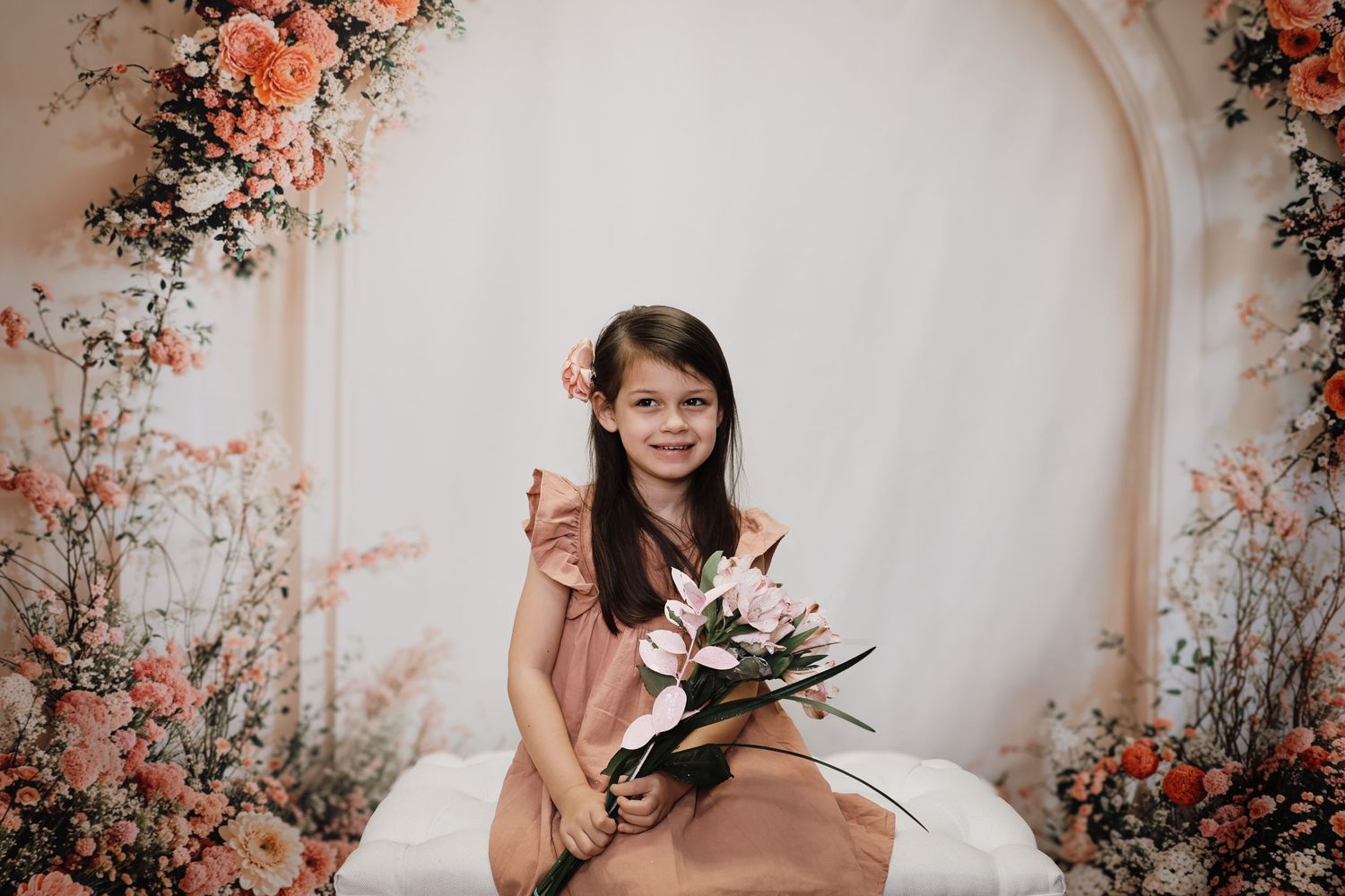 Child in pink dress holds floral bouquet while sitting against elegant archway decorated with coral and peach flowers.