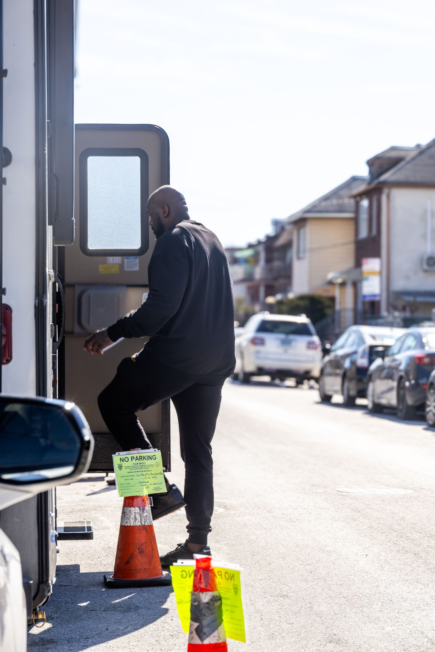 Person in dark clothing using an ATM machine on a suburban street with traffic cones and parked cars in the background.