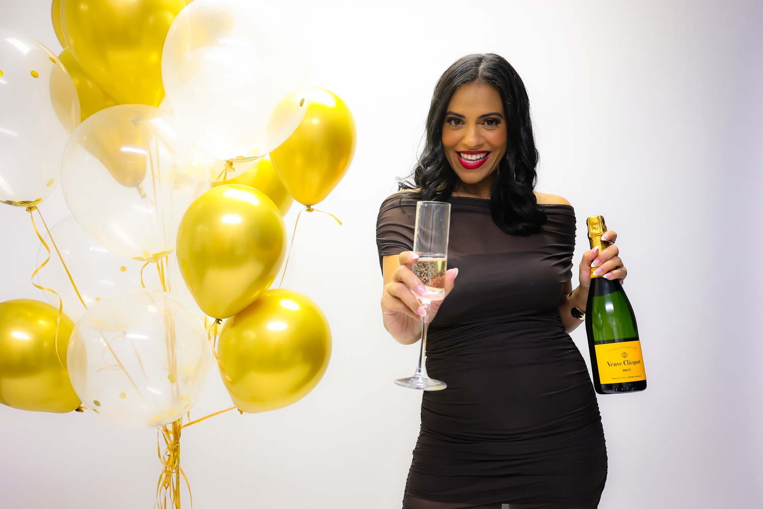 Woman in black dress holding champagne bottle and glass celebrating with gold balloons in background.