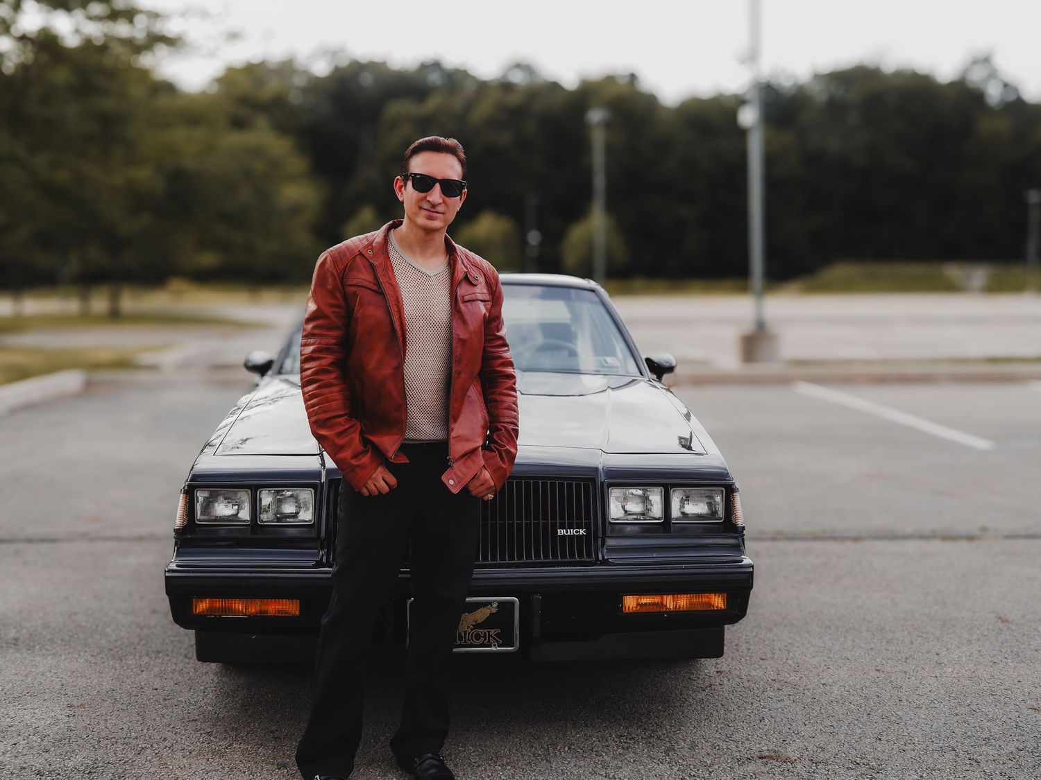 A person in a brown leather jacket poses with a classic black Buick Grand National car in a parking lot.