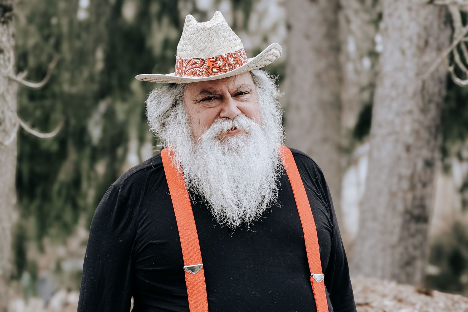 An elderly man with a long white beard wearing orange suspenders and a black shirt stands in a wooded outdoor setting.
