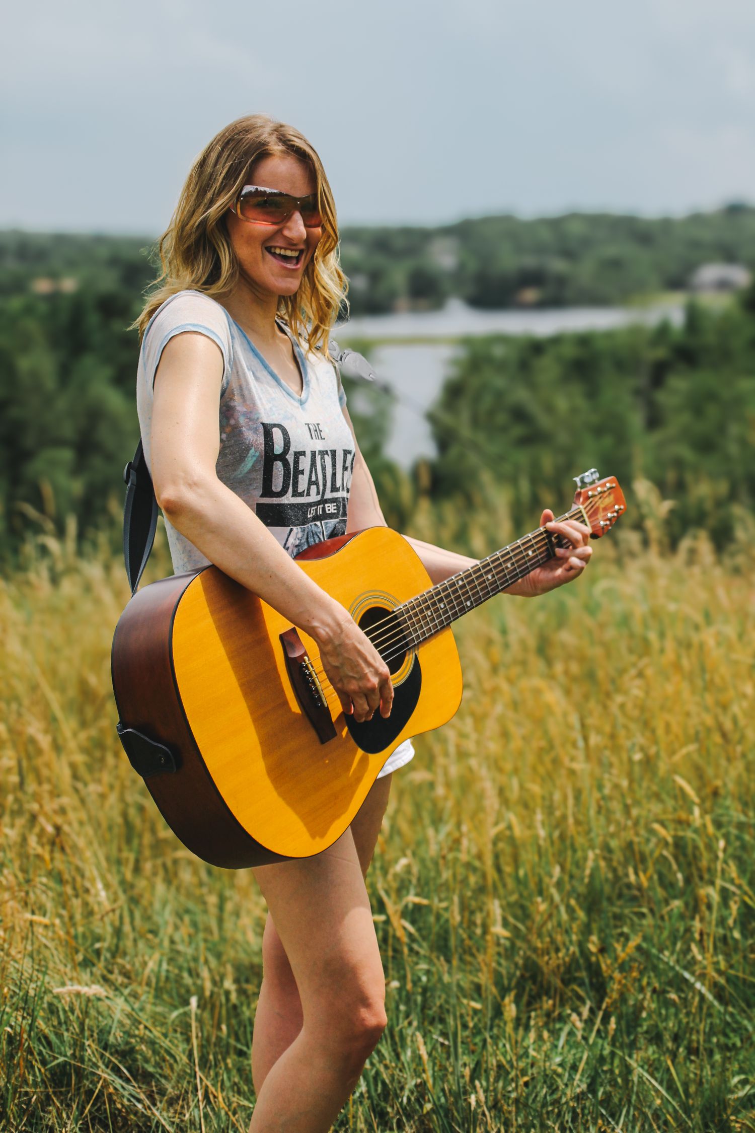 A musician plays an acoustic guitar while standing in a sunlit field of tall grass and wildflowers.