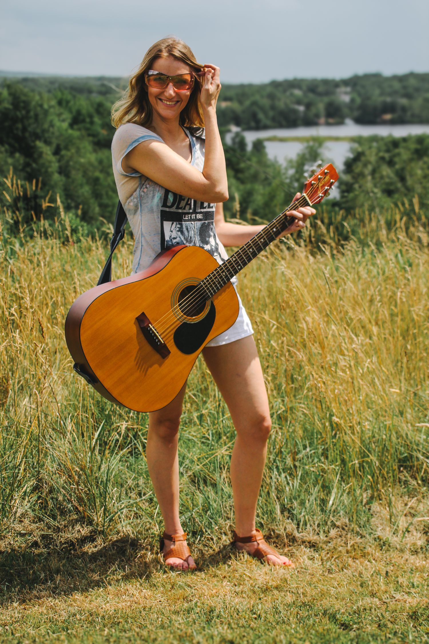 A musician poses with an acoustic guitar in a sunny meadow overlooking a lake and green hills.