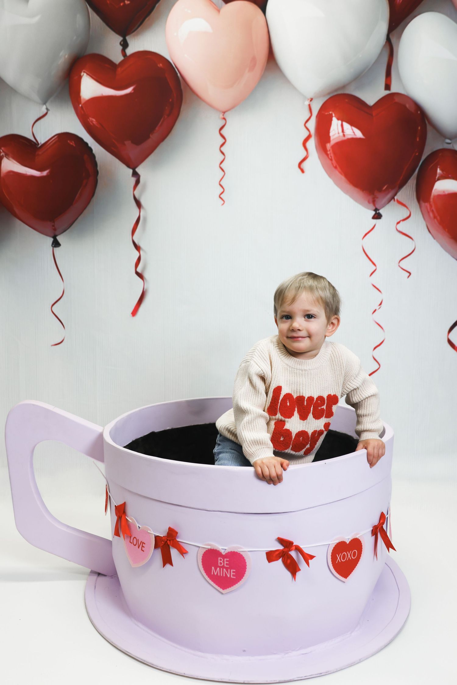 Valentine's Day photo setup with heart balloons, purple curved backdrop and festive decorations.