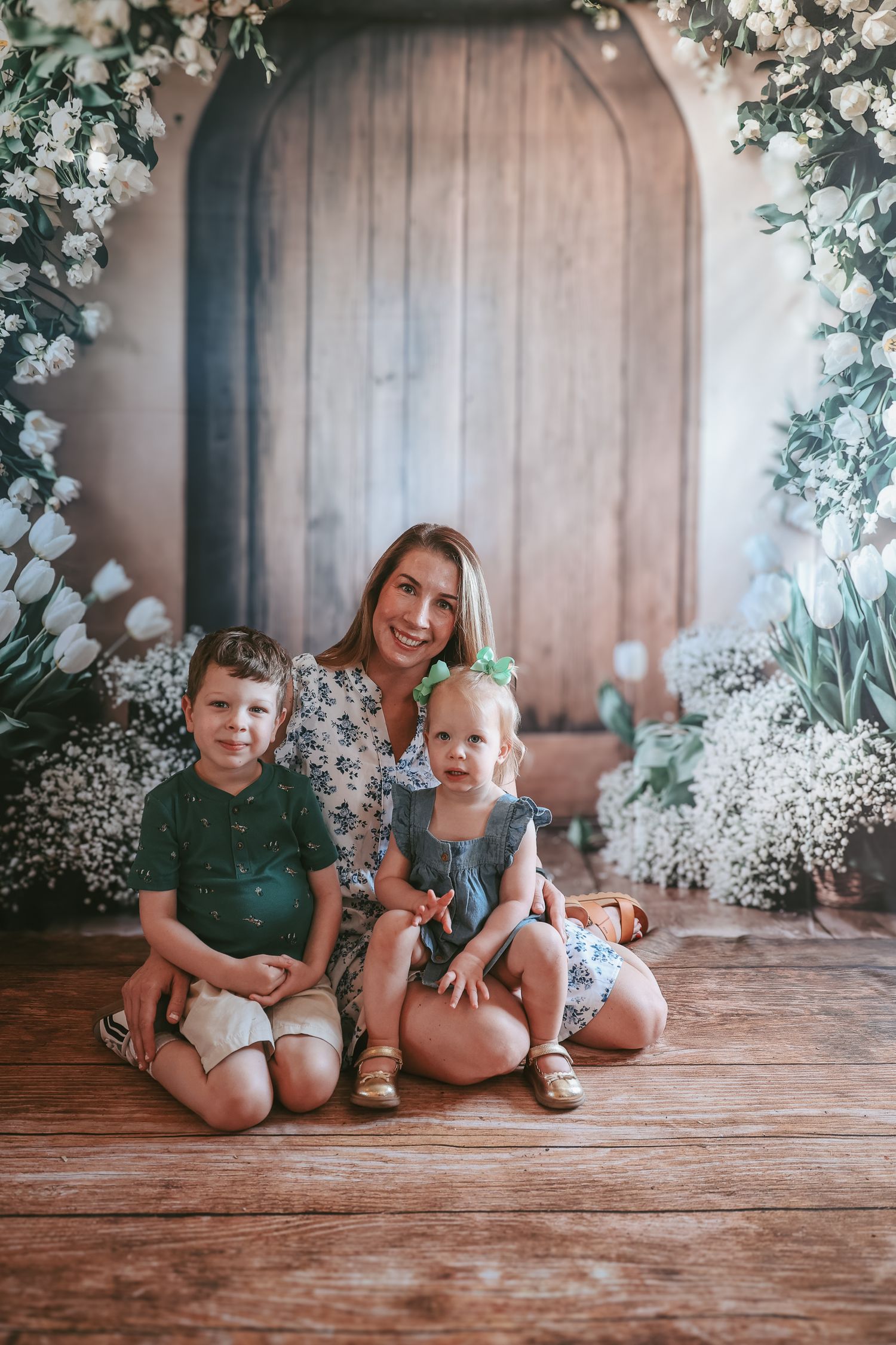Family portrait with floral arch backdrop and baby's breath flowers on rustic wooden floor in soft lighting.