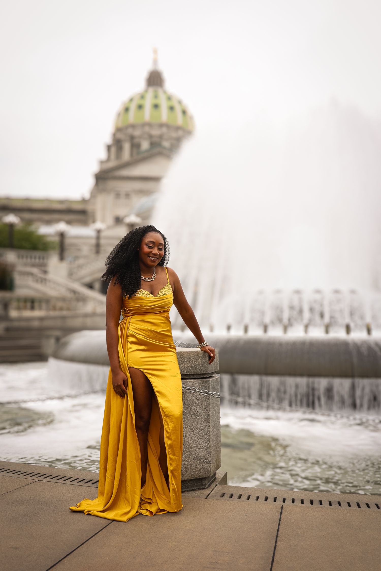 A woman in a flowing yellow dress poses gracefully by a fountain at a grand architectural landmark.