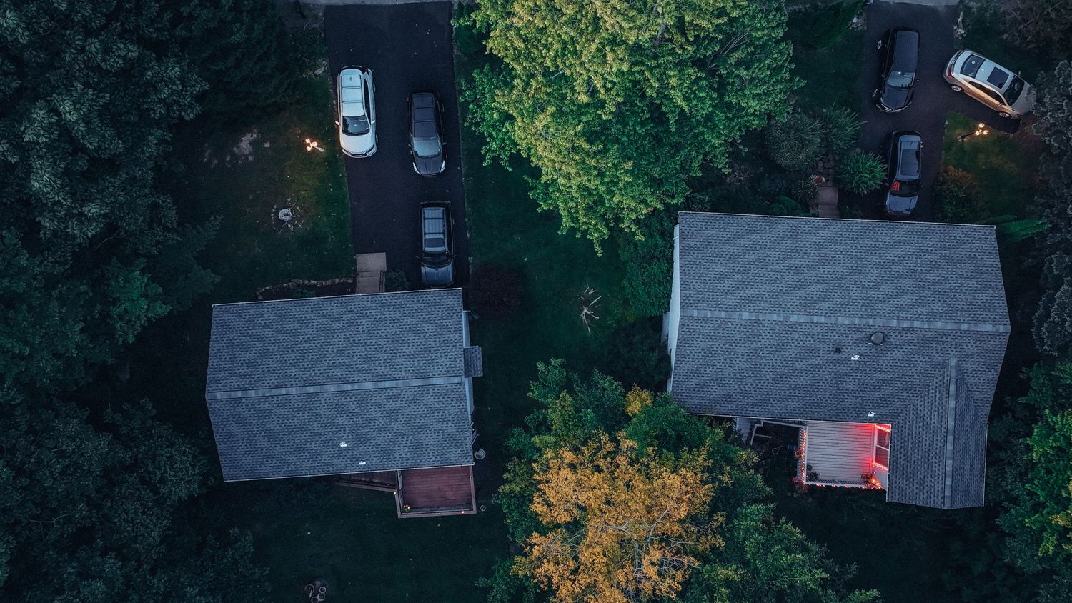 Aerial drone view of two suburban houses with gray roofs surrounded by lush green trees at dusk.