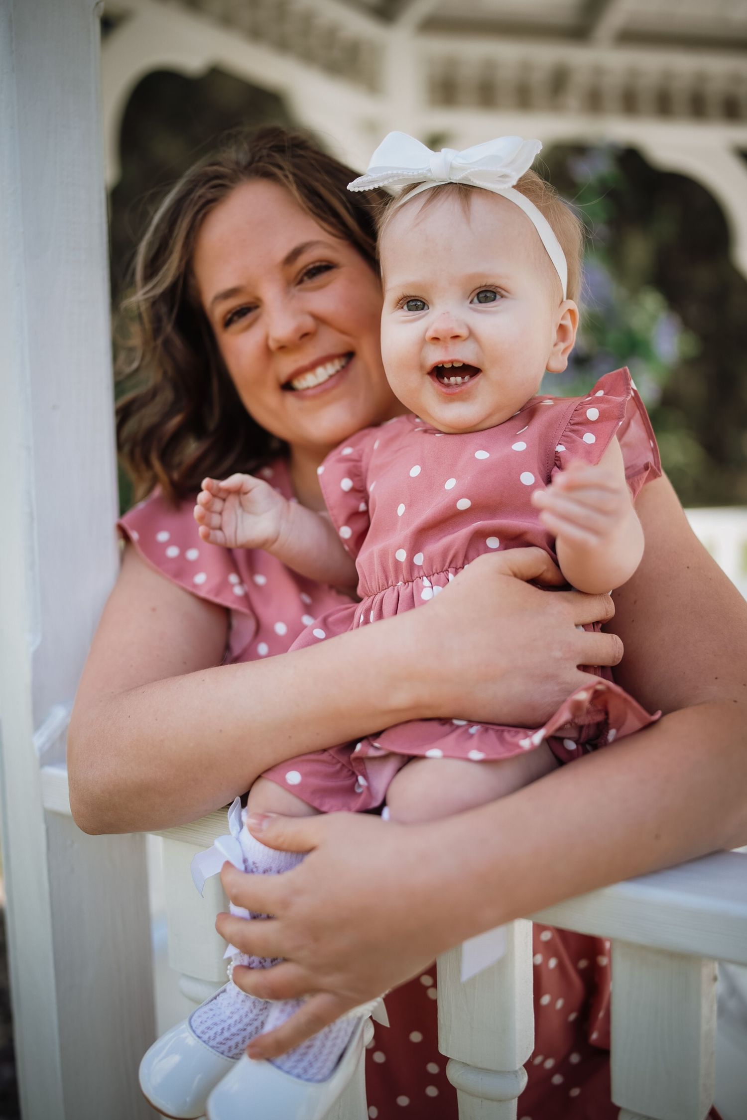 A smiling mother holds her happy baby girl, both wearing matching pink polka dot dresses near a white gazebo.