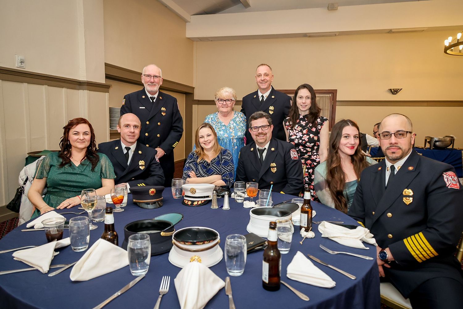 Group of people in formal attire and fire department uniforms posing together at a banquet dinner table with navy blue tablecloth.