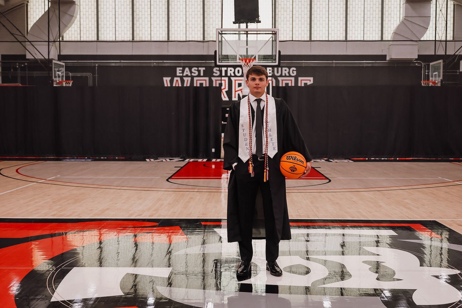 Graduate in cap and gown holds basketball on East Stroudsburg University court during senior photo session.