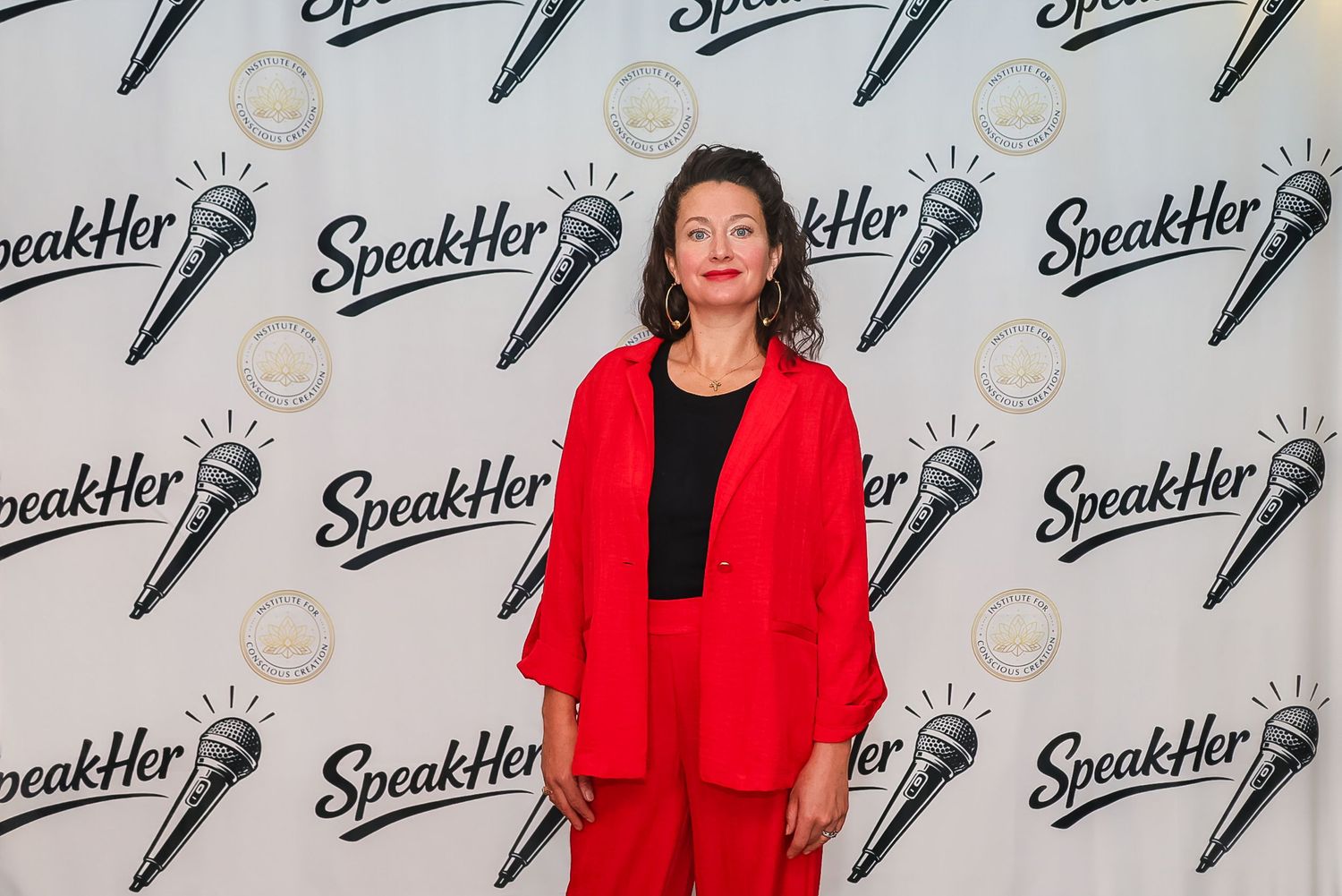 Woman in red suit posing at SpeakHer event backdrop with microphone logos.