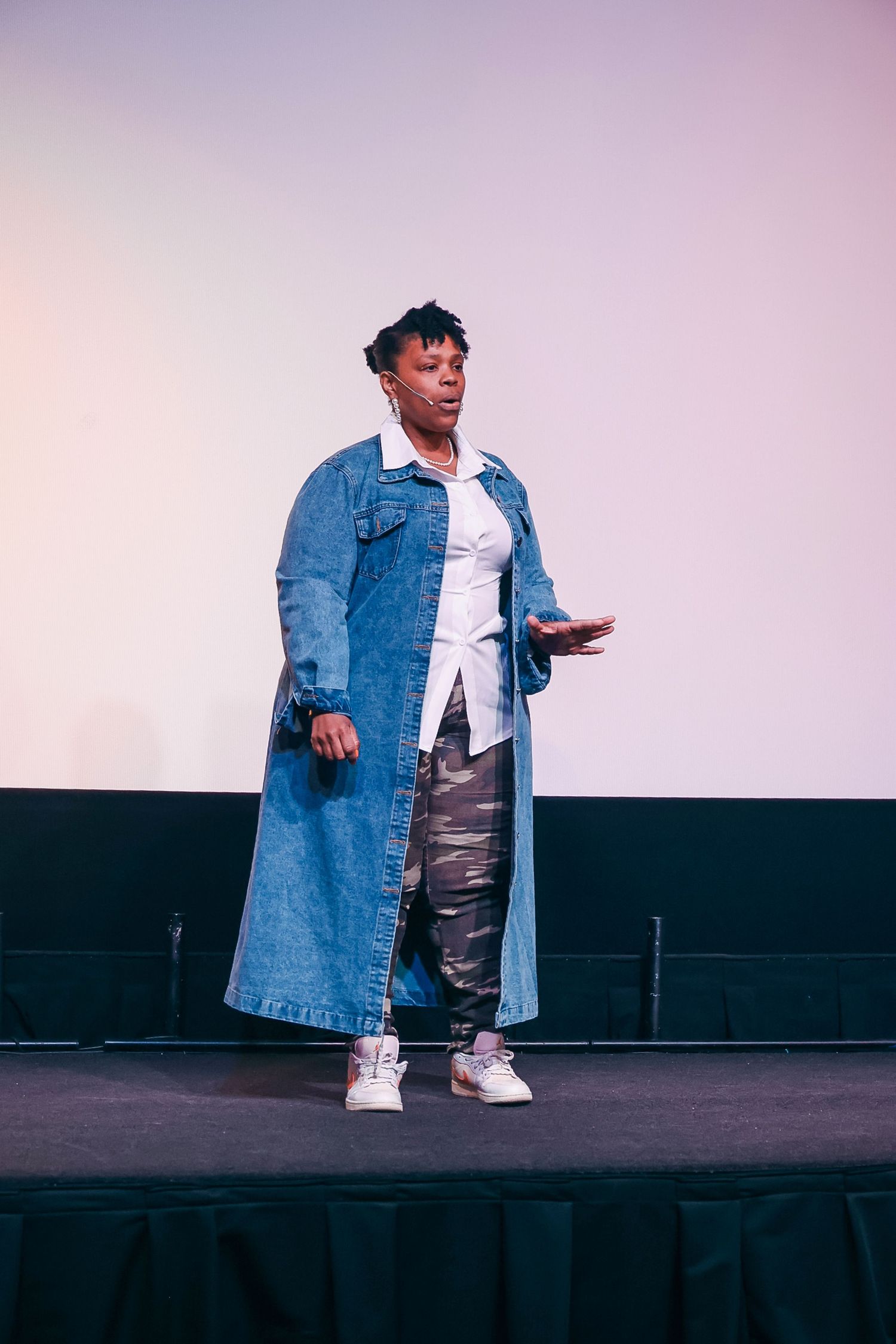 Person on stage wearing a long denim coat, white shirt, camo pants, and white sneakers, gesturing while speaking.