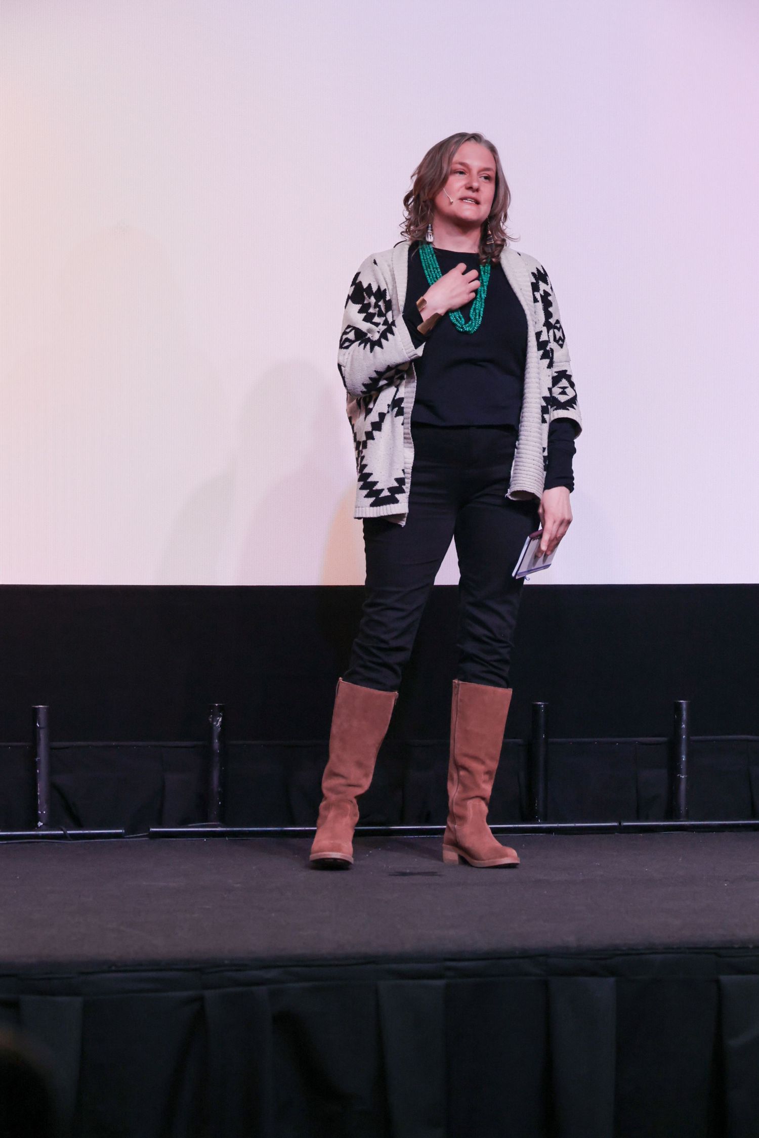 Woman in patterned cardigan, black pants, and brown boots presenting on stage with a green necklace.