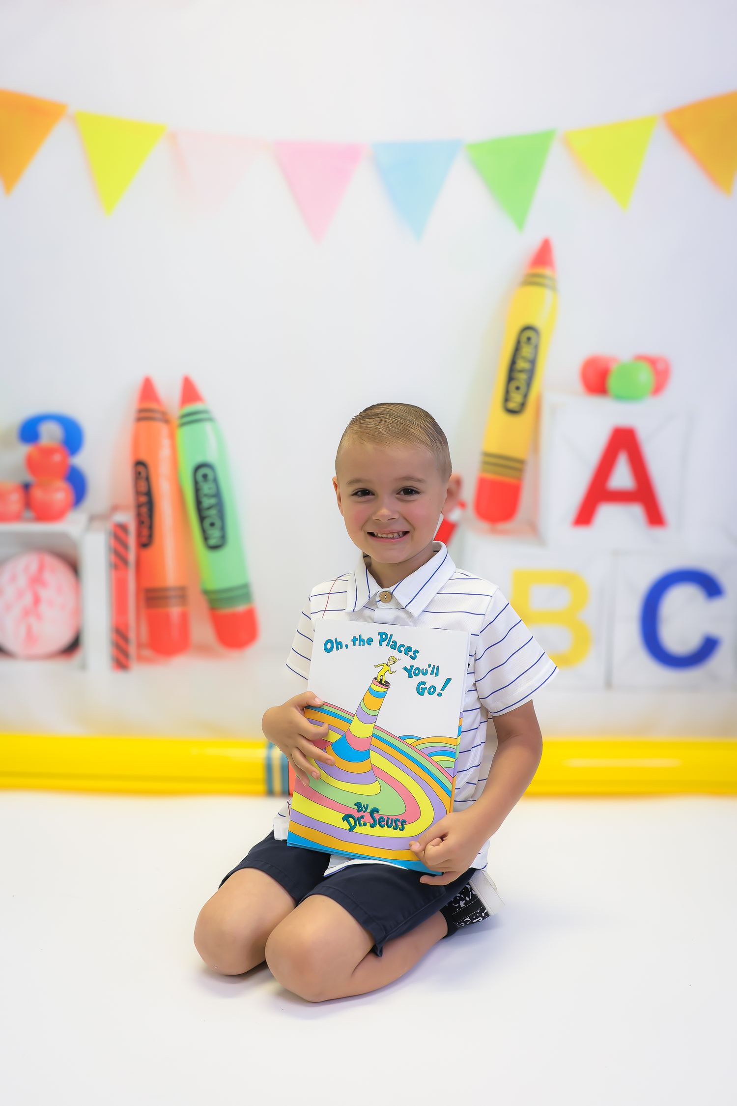 A smiling young boy sits on the floor holding a Dr. Seuss book, surrounded by colorful crayon decorations and ABC letters.