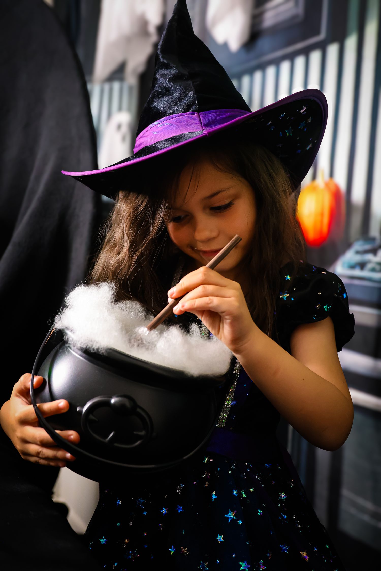 Girl in witch costume with purple hat stirs a smoking cauldron with a wand on Halloween.