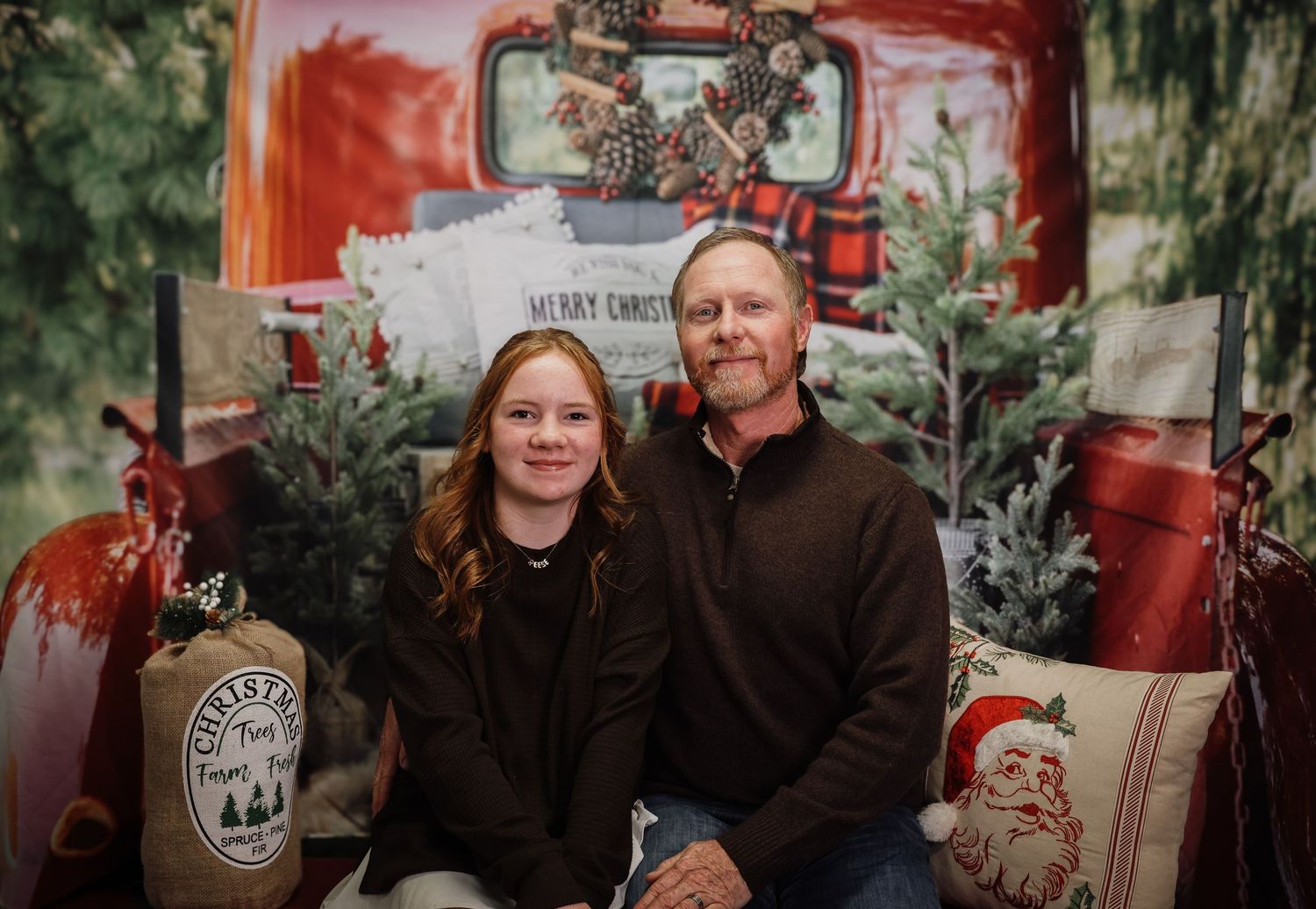 A young girl and middle-aged man pose together in front of a festive Christmas backdrop with a red truck and holiday decor.