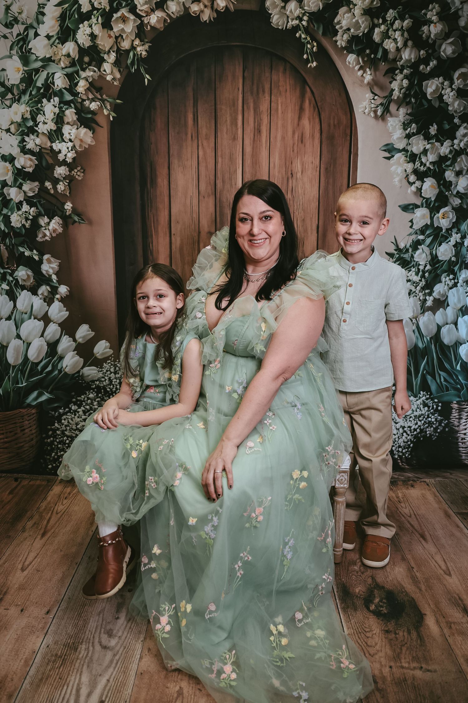 A woman in a green floral gown poses with two children before a wooden door adorned with white flowers.