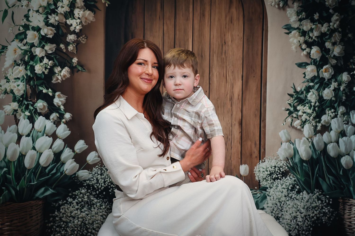 A woman in white dress holds a young boy on her lap, surrounded by white tulips and floral decor in a studio portrait.