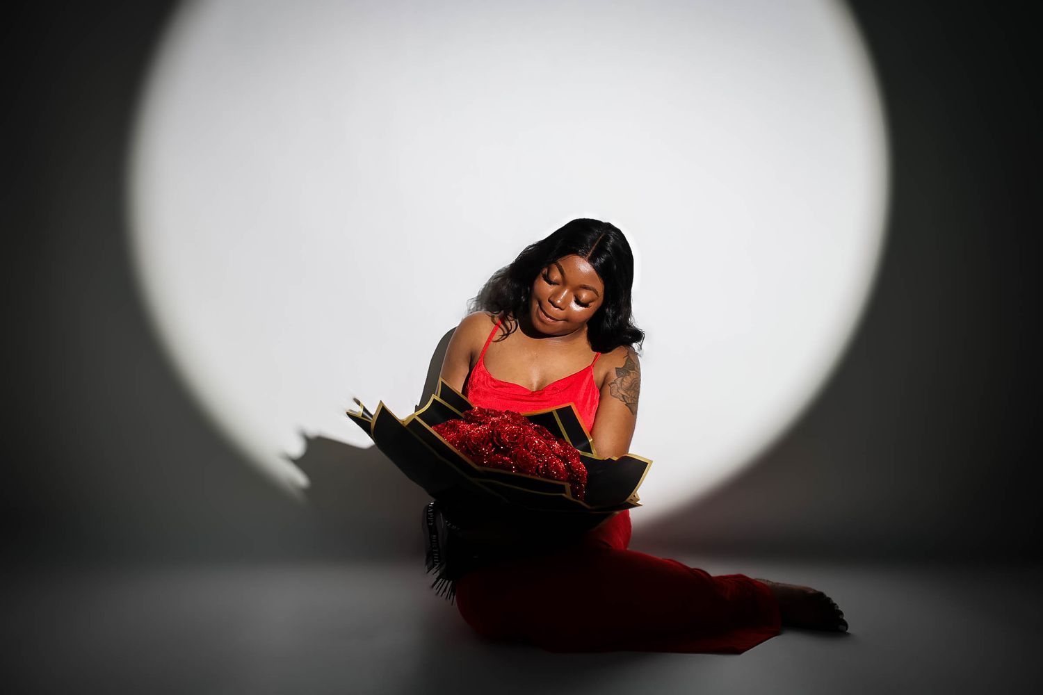 Woman in red dress sitting on floor reading a book, illuminated by a circular spotlight against a gray background.