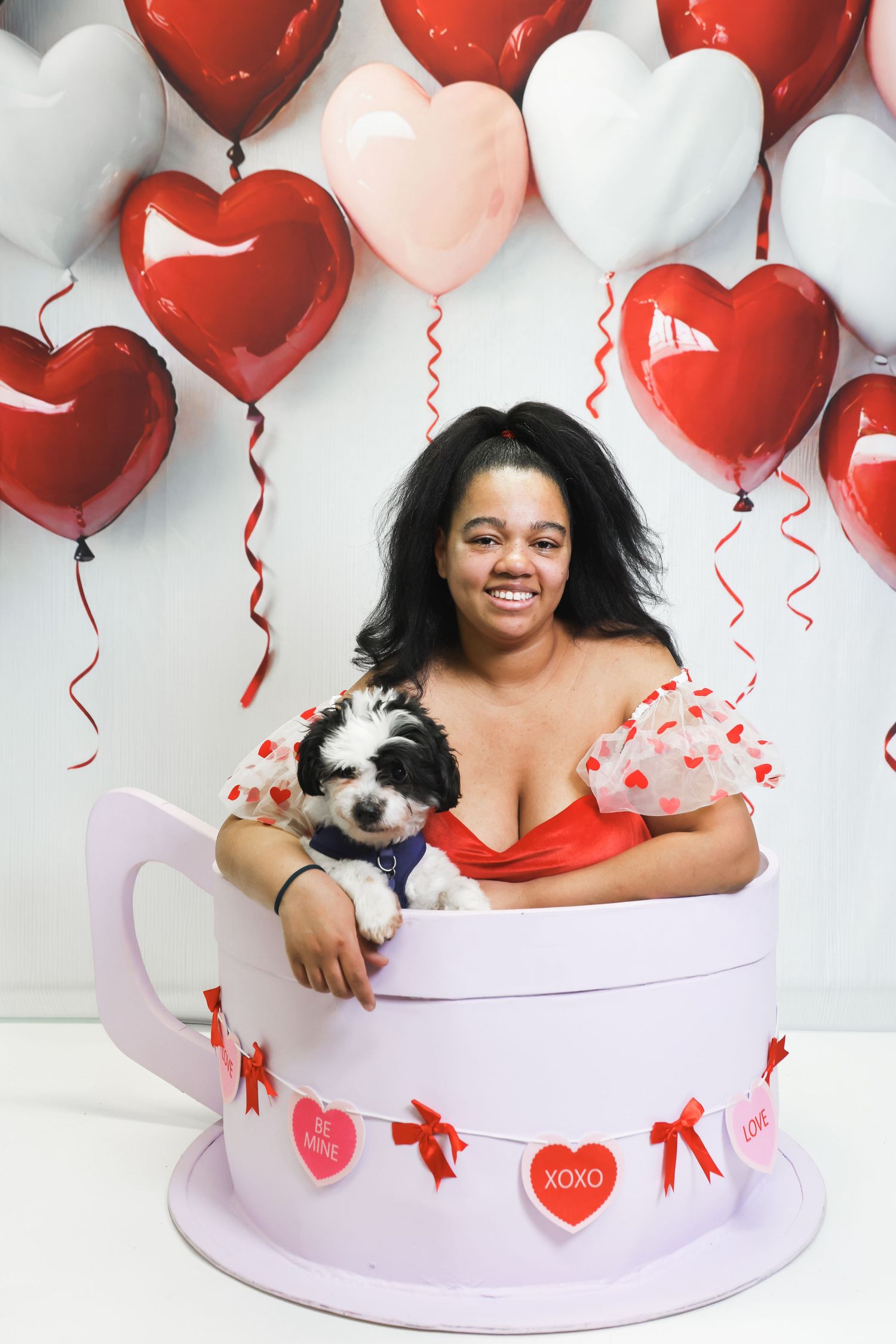 Woman smiling in giant teacup prop with puppy, surrounded by red and pink heart balloons for Valentine's Day.