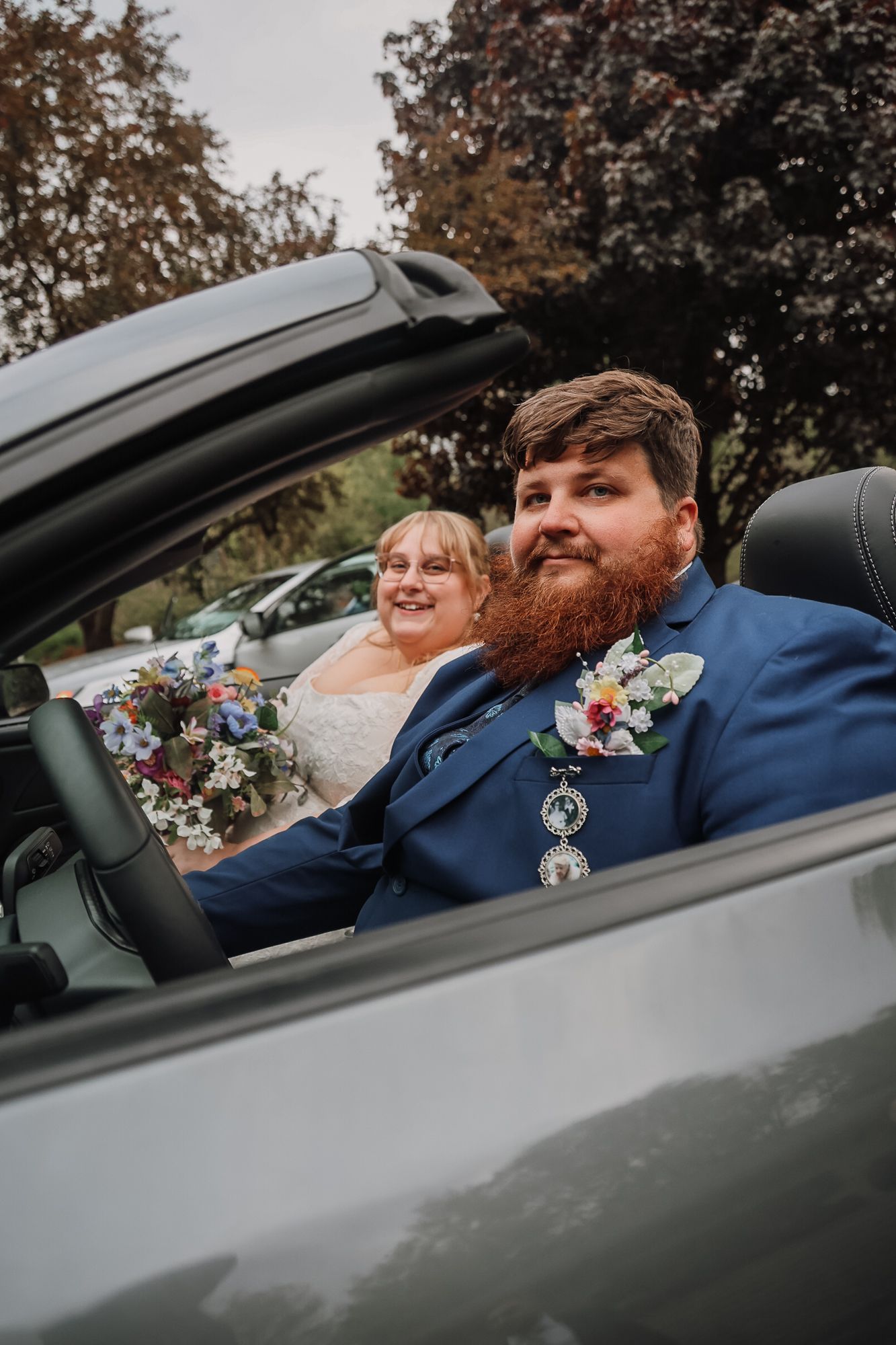 A groom with a red beard in a blue suit and bride in white holding flowers sit in a convertible car on their wedding day.