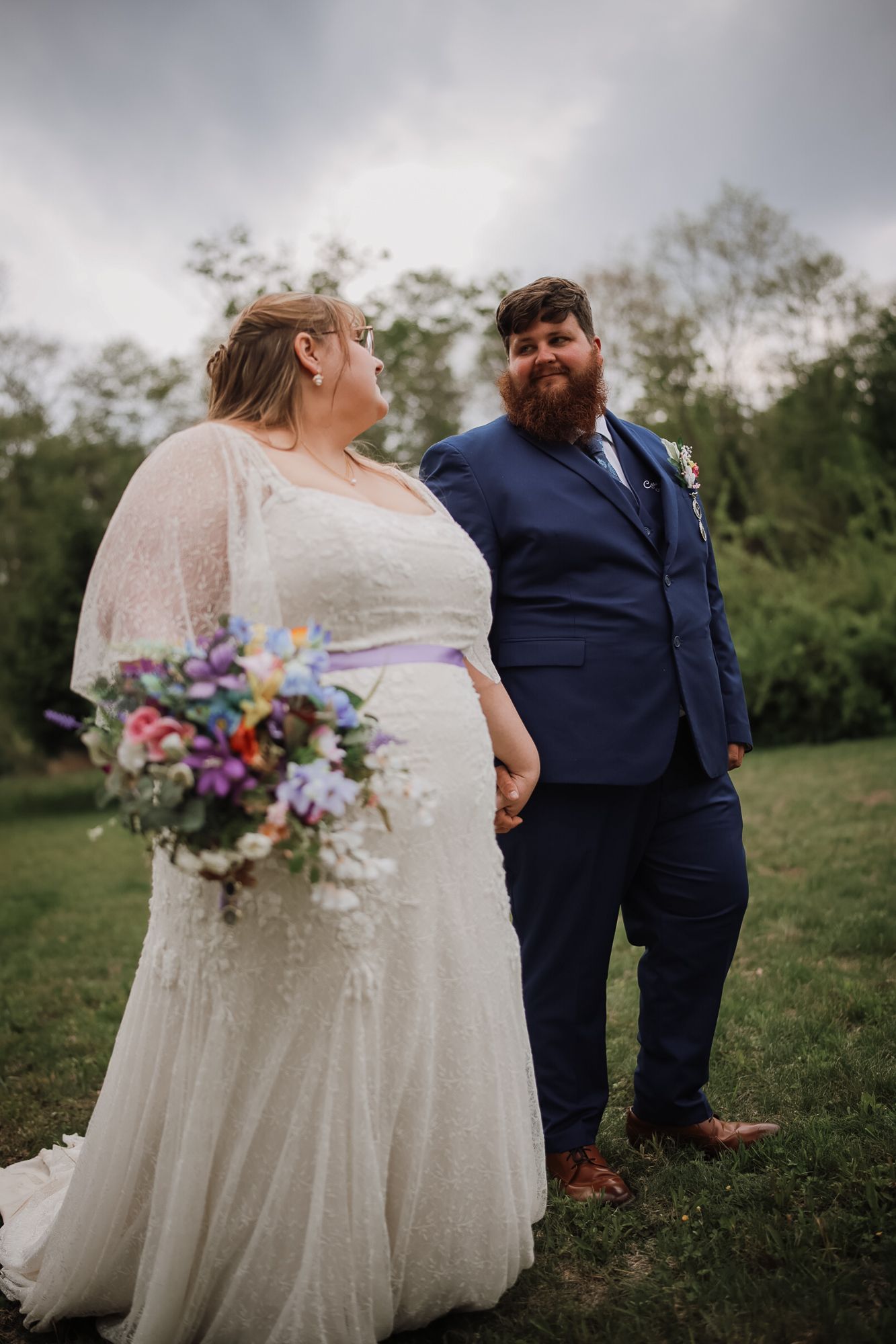 Bride in lace wedding gown holding colorful bouquet gazes at bearded groom in navy suit on green lawn.