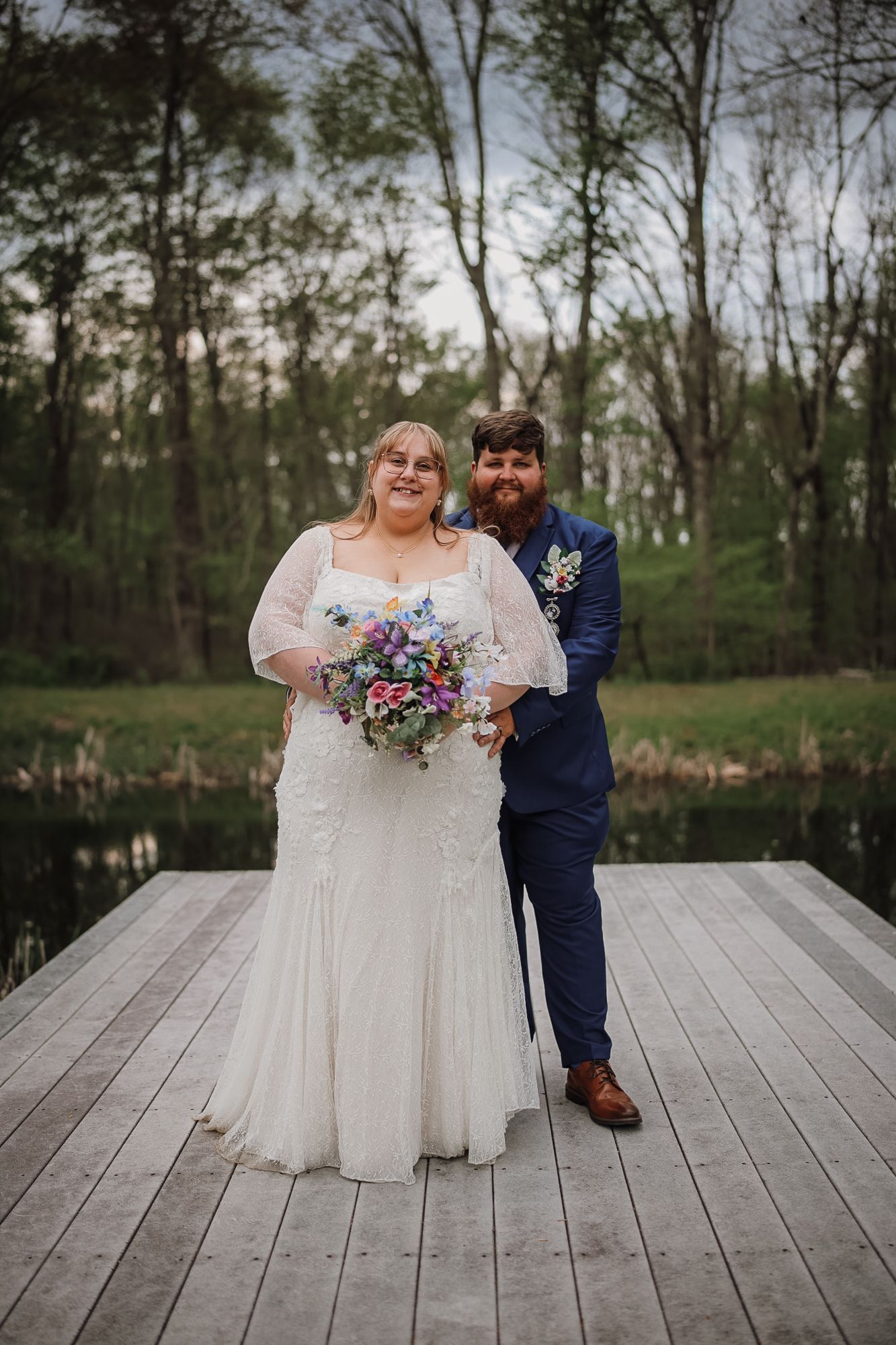 Bride and groom pose on a wooden dock surrounded by trees and a pond, bride holding a colorful wildflower bouquet.