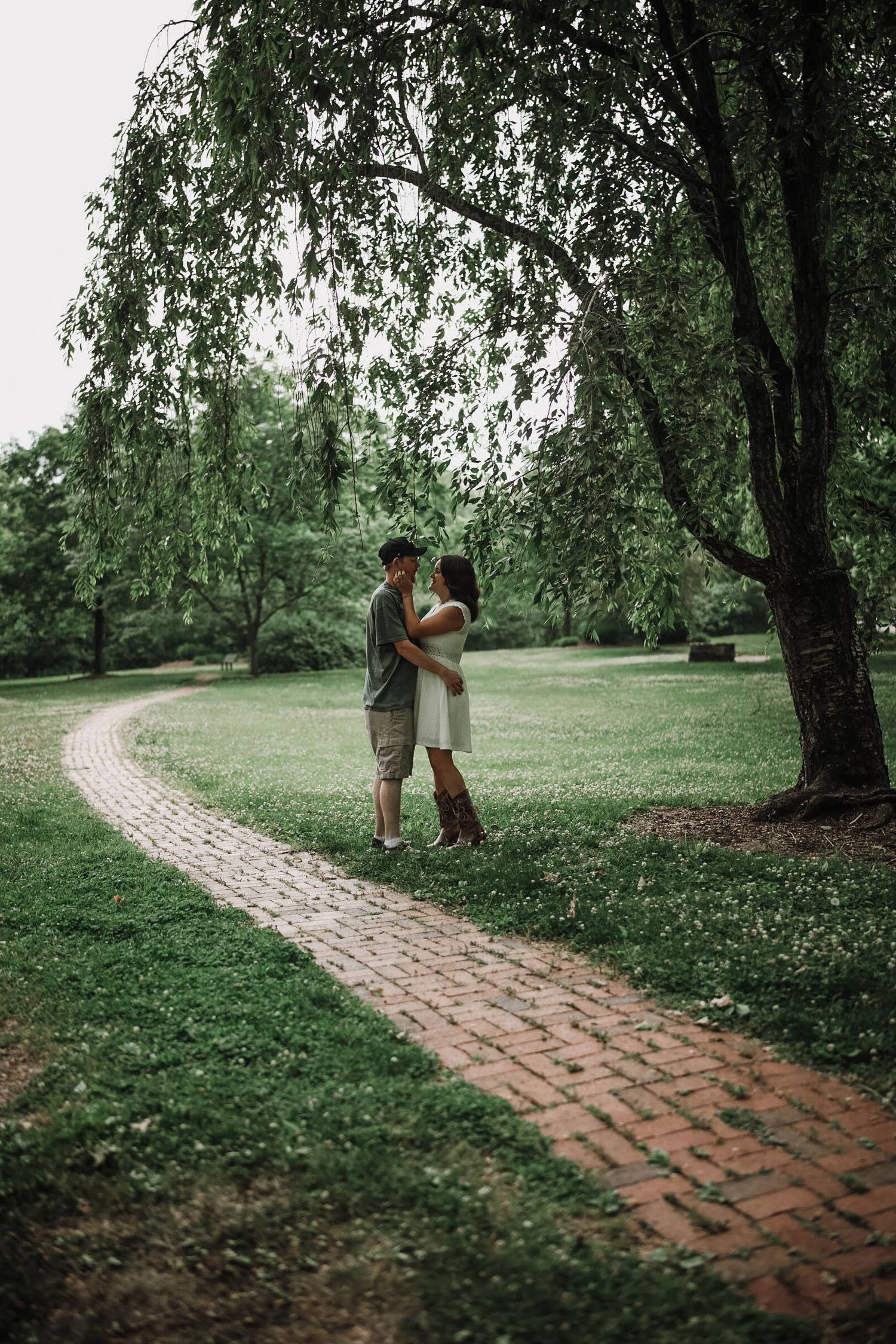 Couple embracing on a brick path in a lush green park under a weeping willow tree.