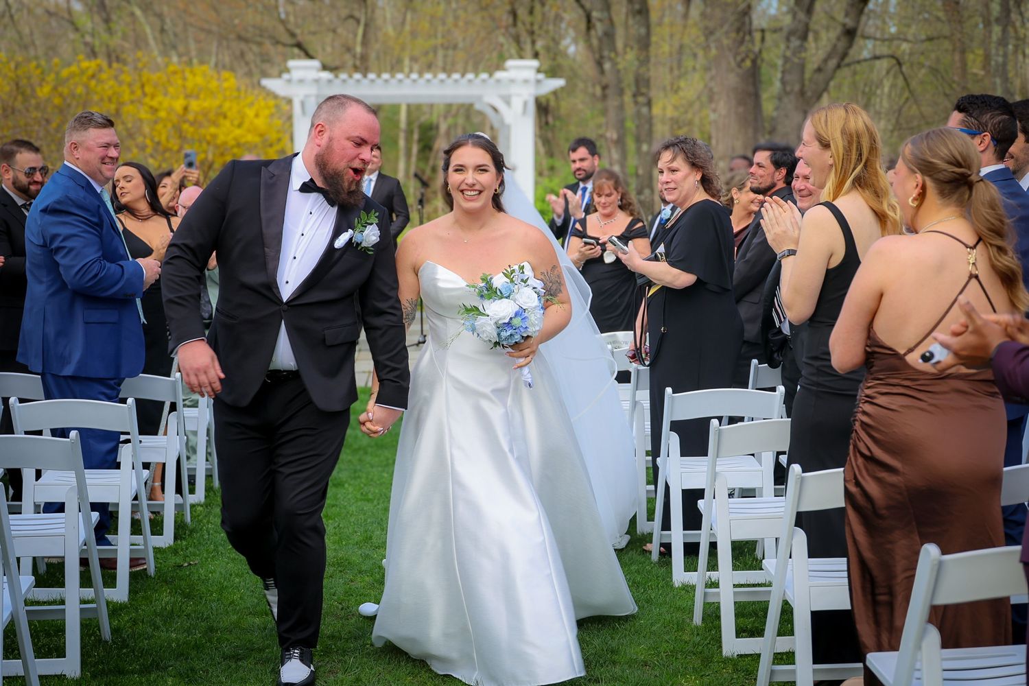Bride and groom walk down outdoor aisle after wedding ceremony, guests applauding, white chairs, floral arch backdrop.