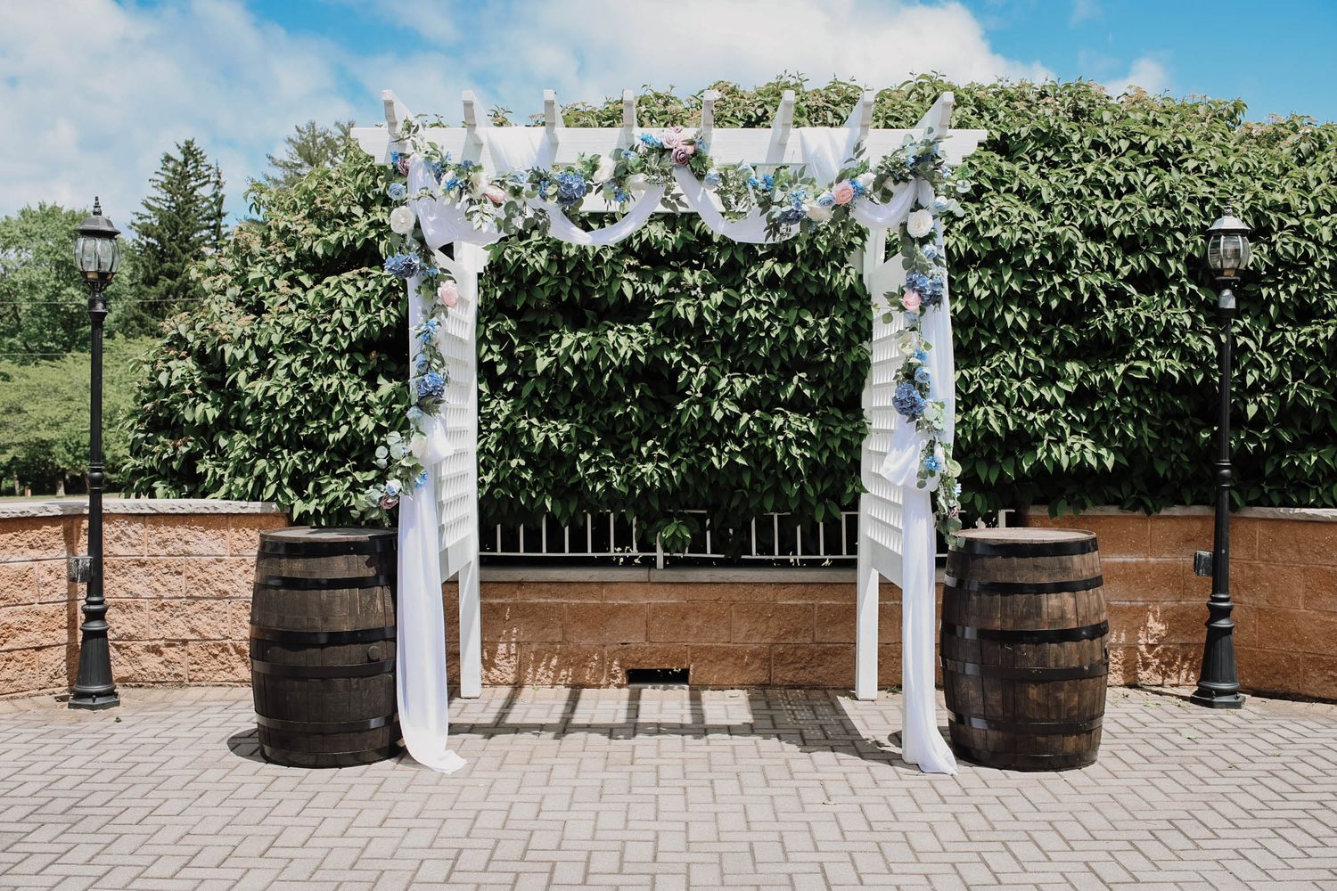 Elegant outdoor wedding arch adorned with white draping and blue flowers, flanked by rustic wooden barrels on a brick patio.