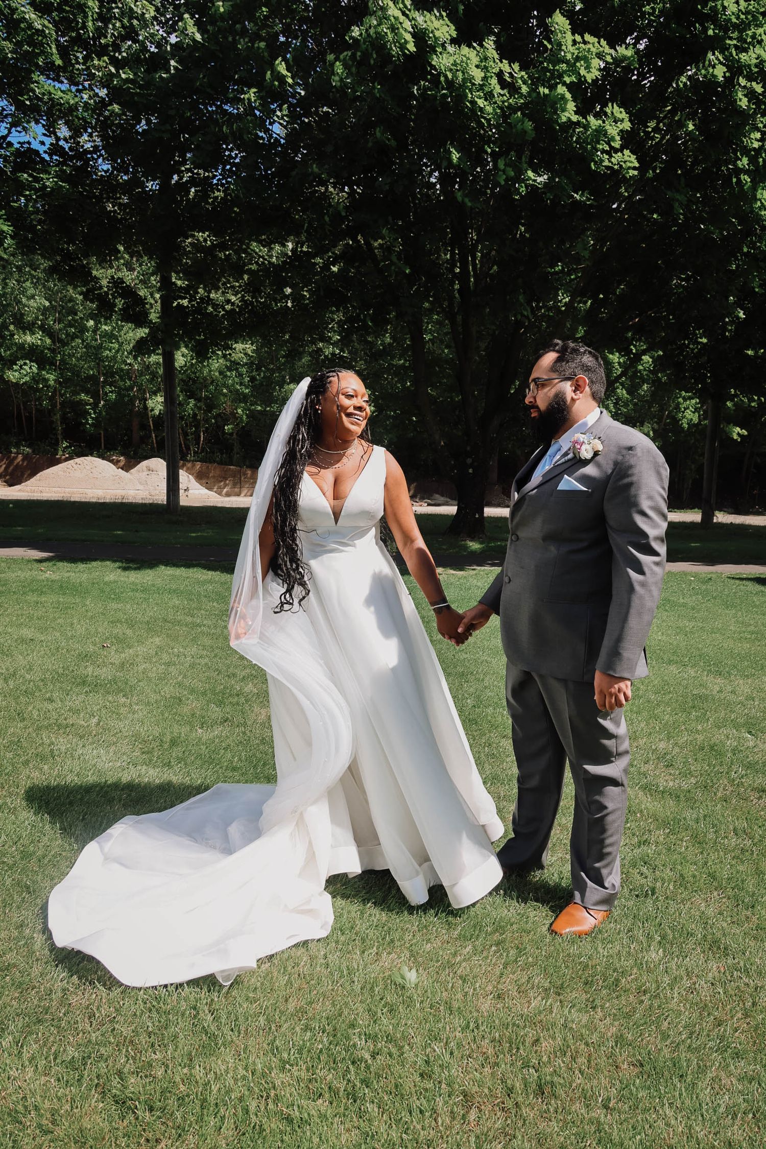 A bride in a white gown and veil holds hands with a groom in a gray suit on a sunny green lawn.