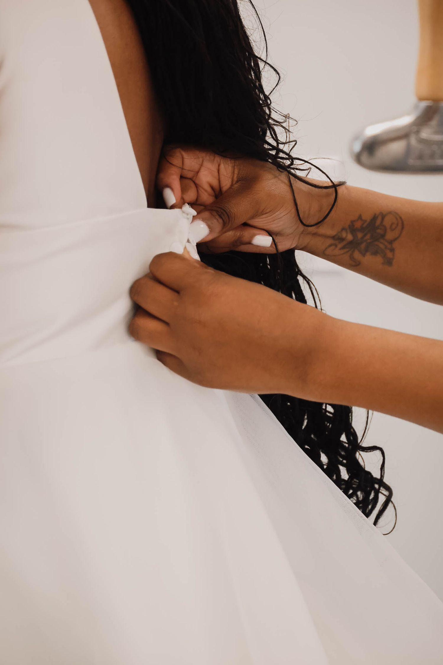 Hands with a floral tattoo fastening the back of a white wedding dress on a bride with long dark hair.