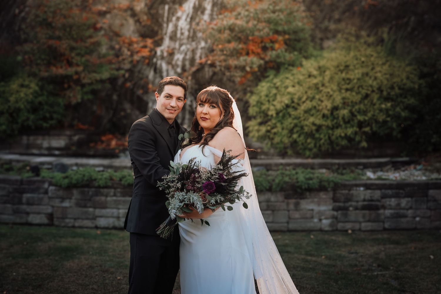Newlywed couple embraces in front of autumn foliage during outdoor wedding photoshoot.