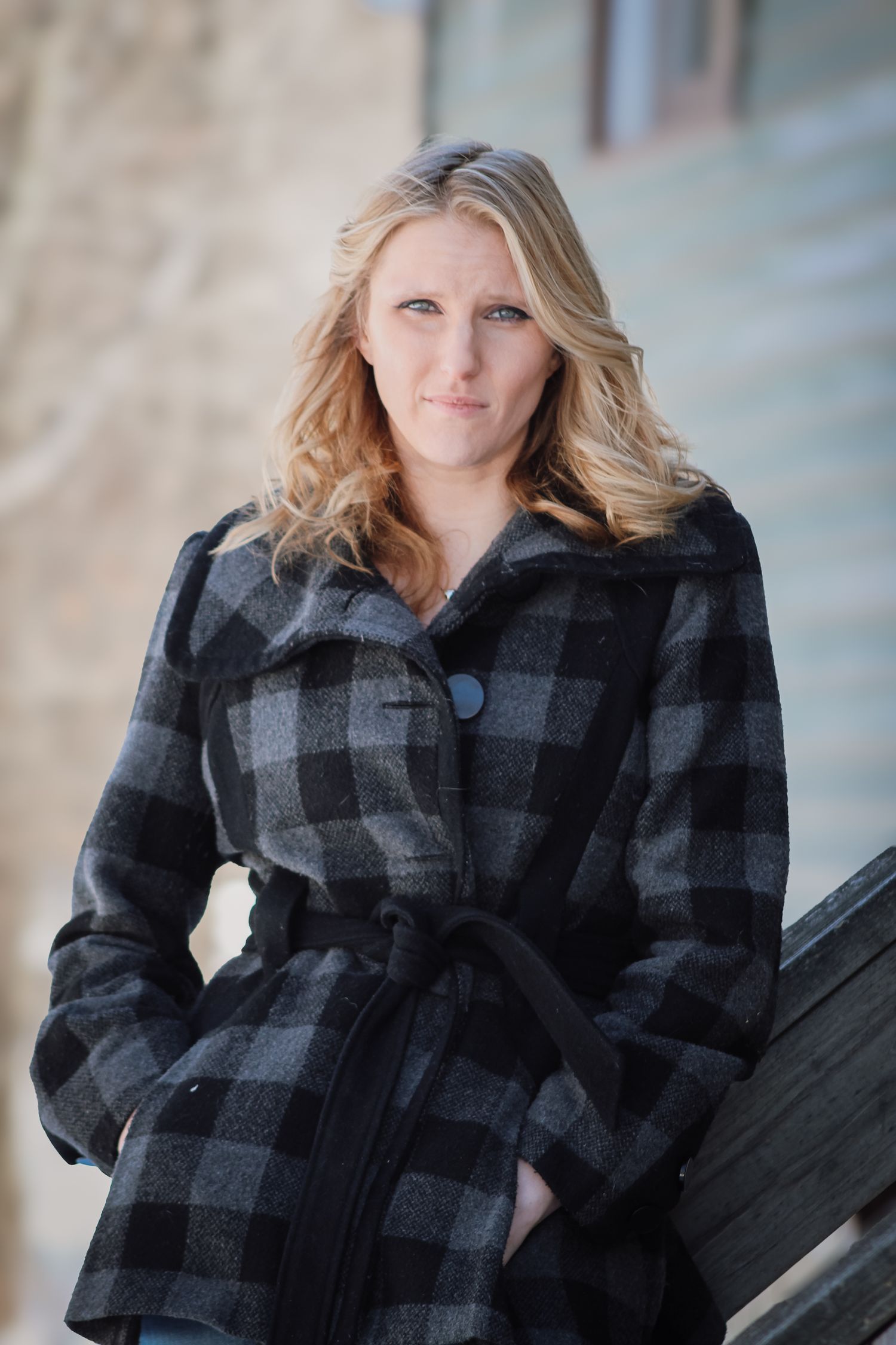 A person wearing a black and gray plaid wool coat with a belt poses outdoors against a light colored wall.