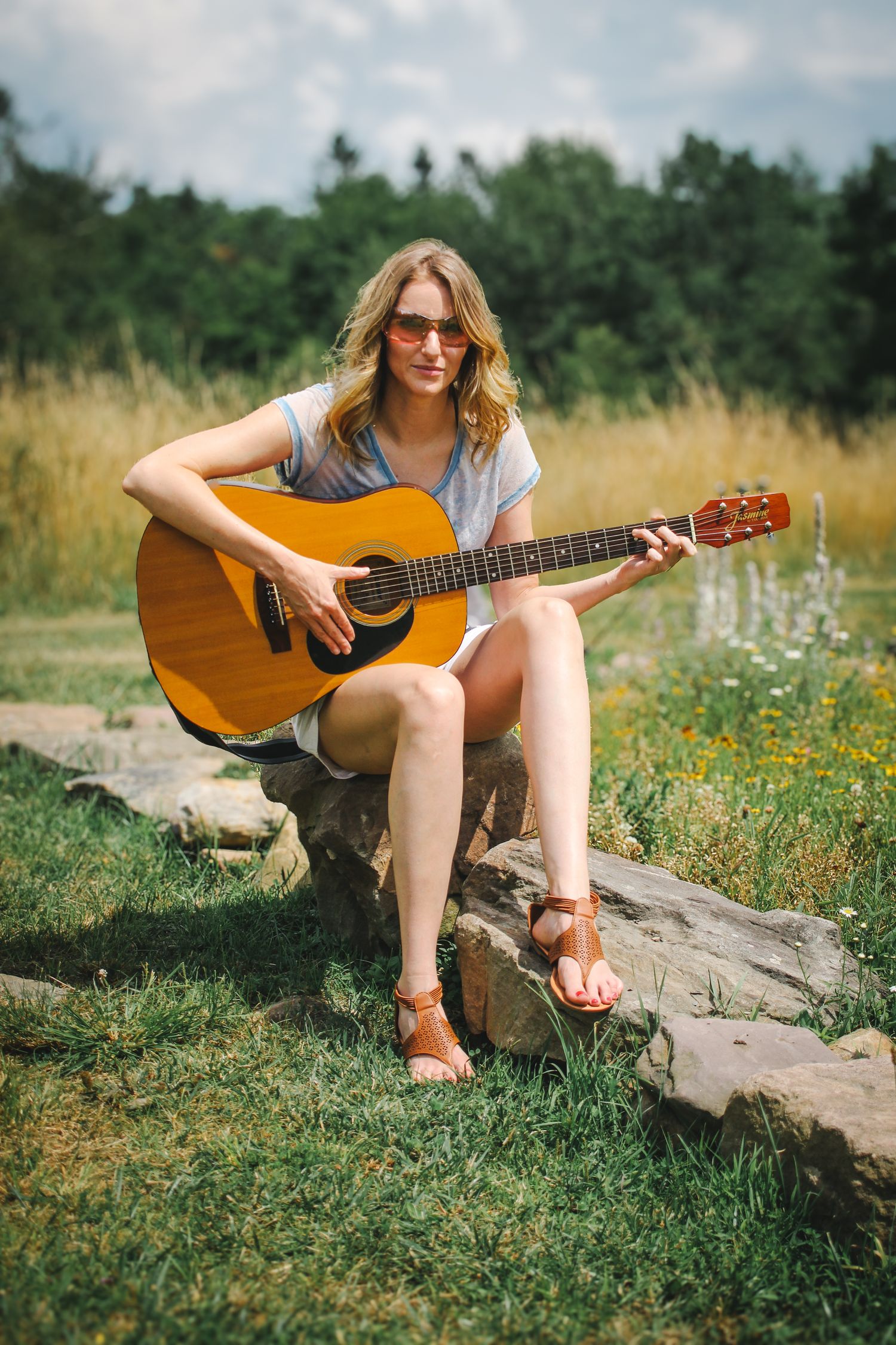 A person wearing sunglasses sits on a rock in a wildflower field playing an acoustic guitar on a sunny summer day.