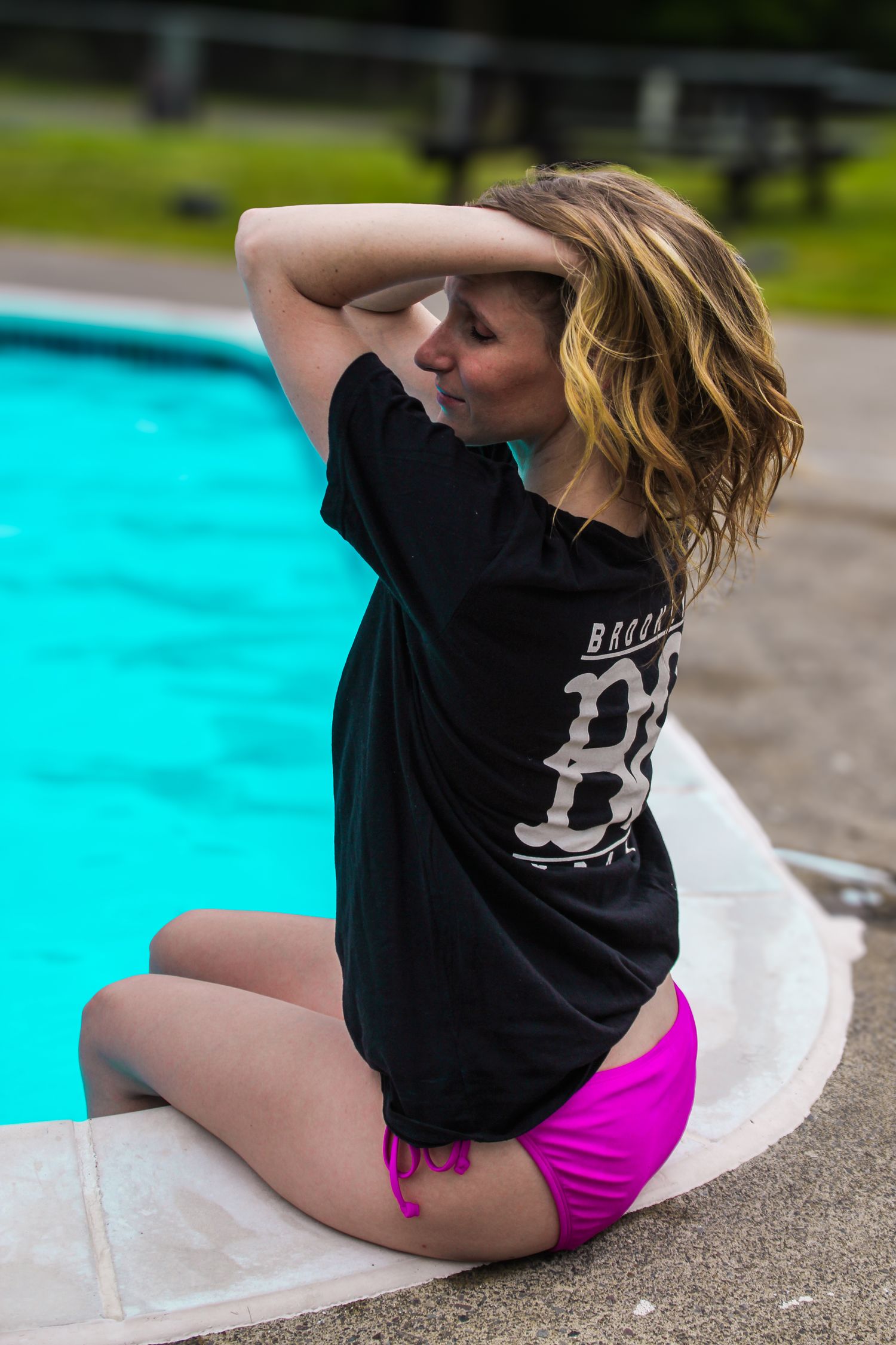 A person in a black t-shirt and pink shorts sits by a pool edge in a casual lounging pose.