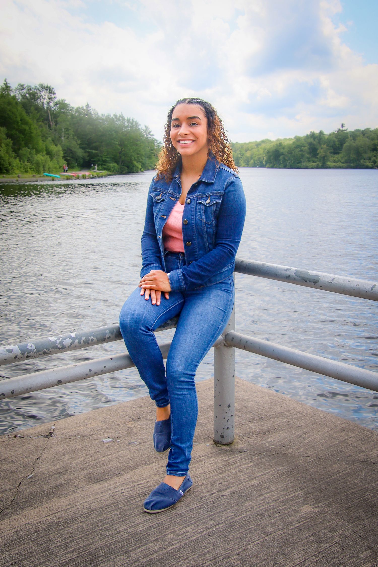 Person in denim jacket sits by lake railing on sunny day with trees in background.